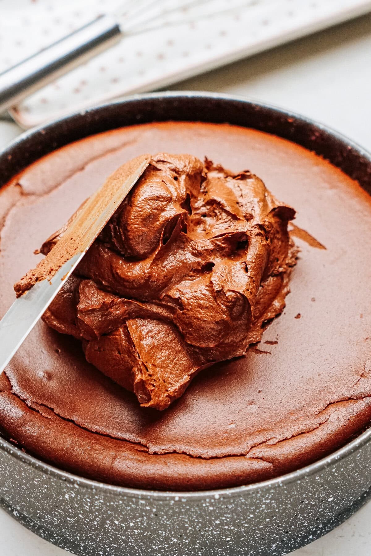 A close-up of chocolate frosting being spread on a round chocolate cake in a baking pan.