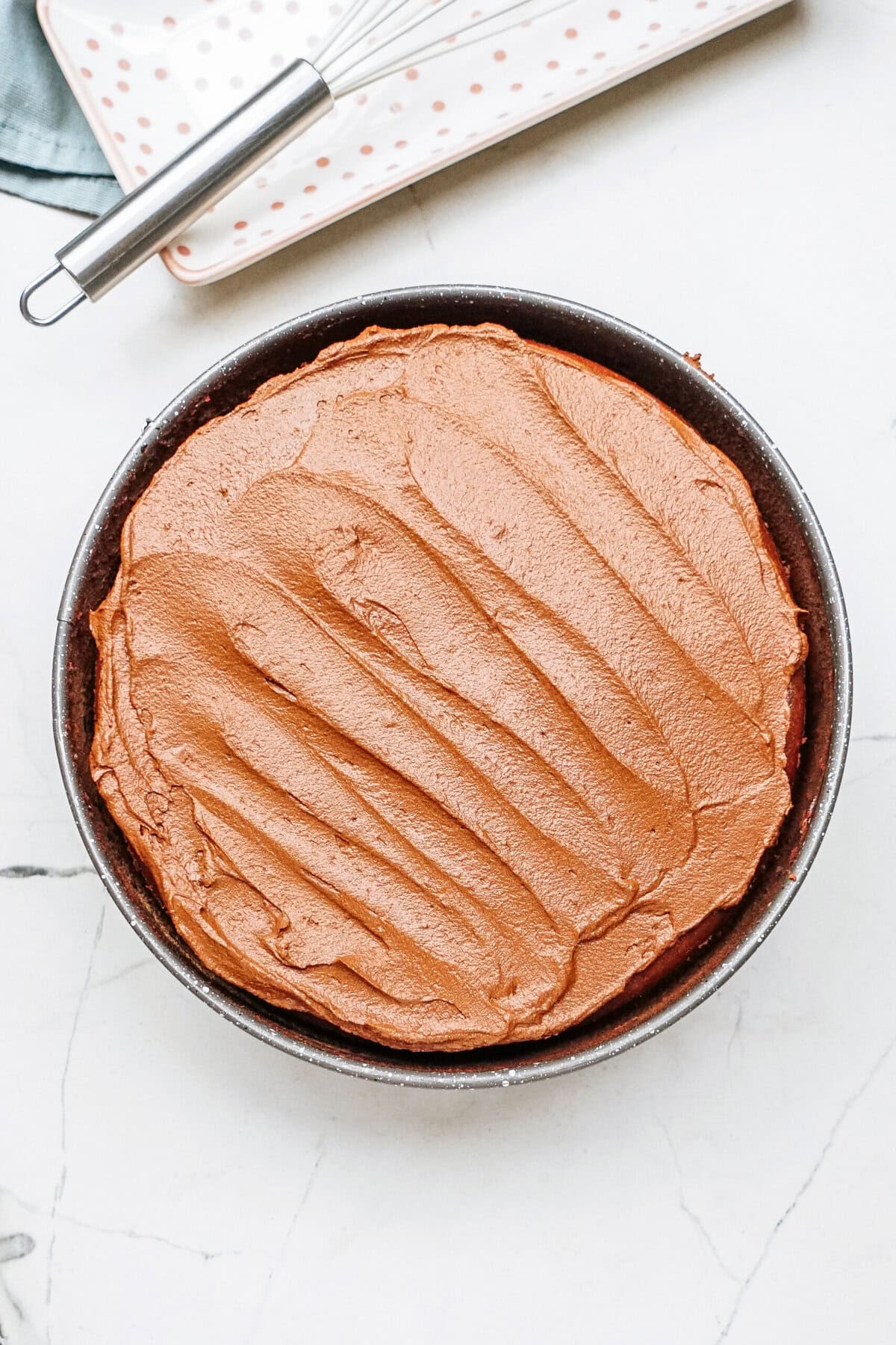 Round cake topped with smooth chocolate frosting in a baking pan on a white marble surface.