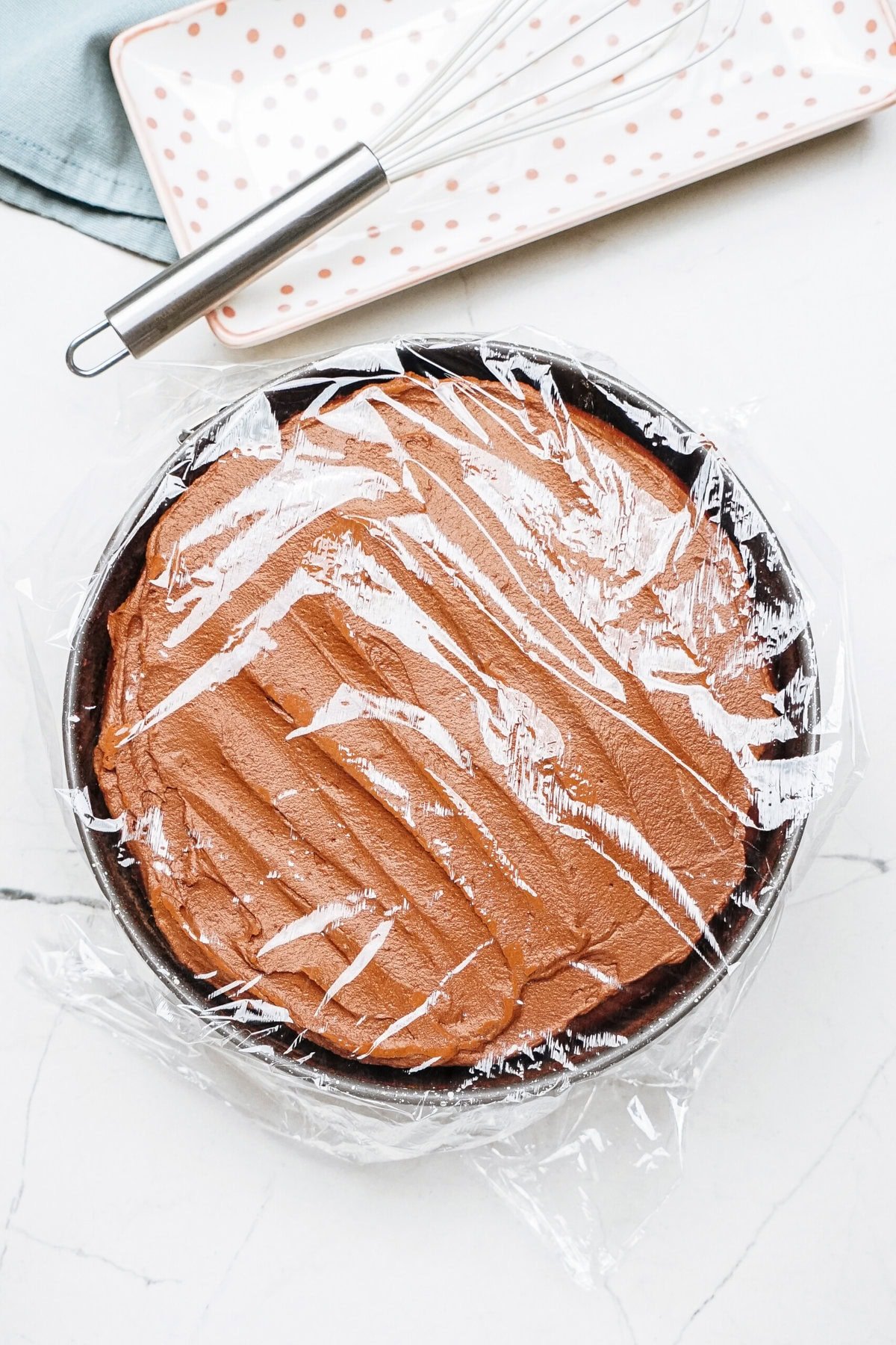 A round chocolate cake covered in plastic wrap on a marble surface, with a whisk and a polka-dot plate in the background.