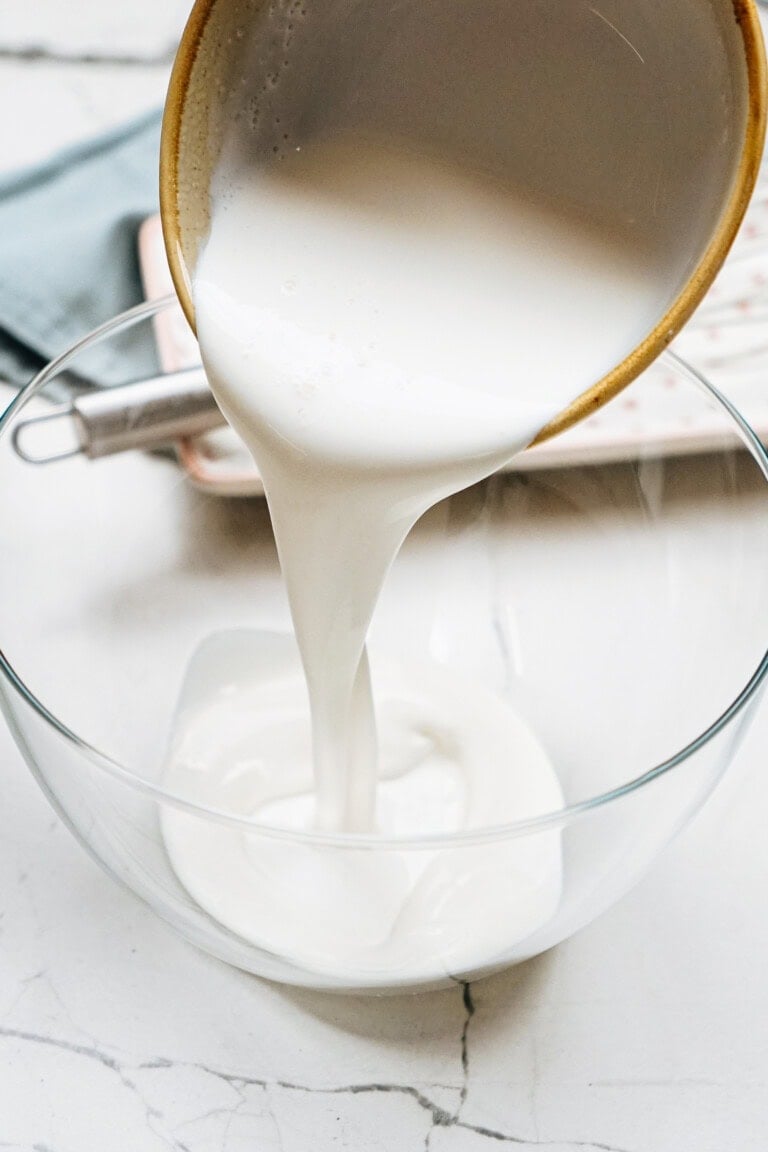 Milk being poured from a ceramic jug into a clear glass bowl on a marble surface.