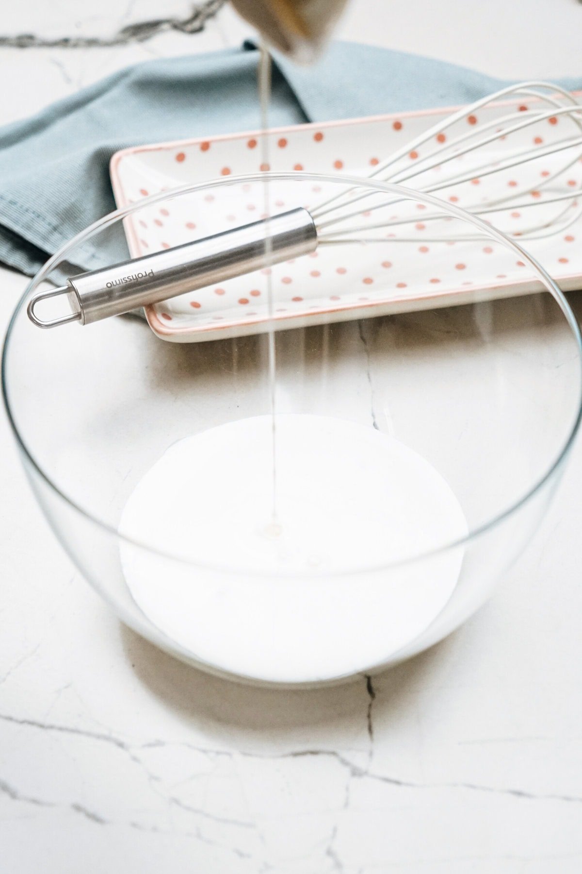 A glass bowl is being filled with liquid. A whisk and a polka dot tray are in the background on a light-colored surface.