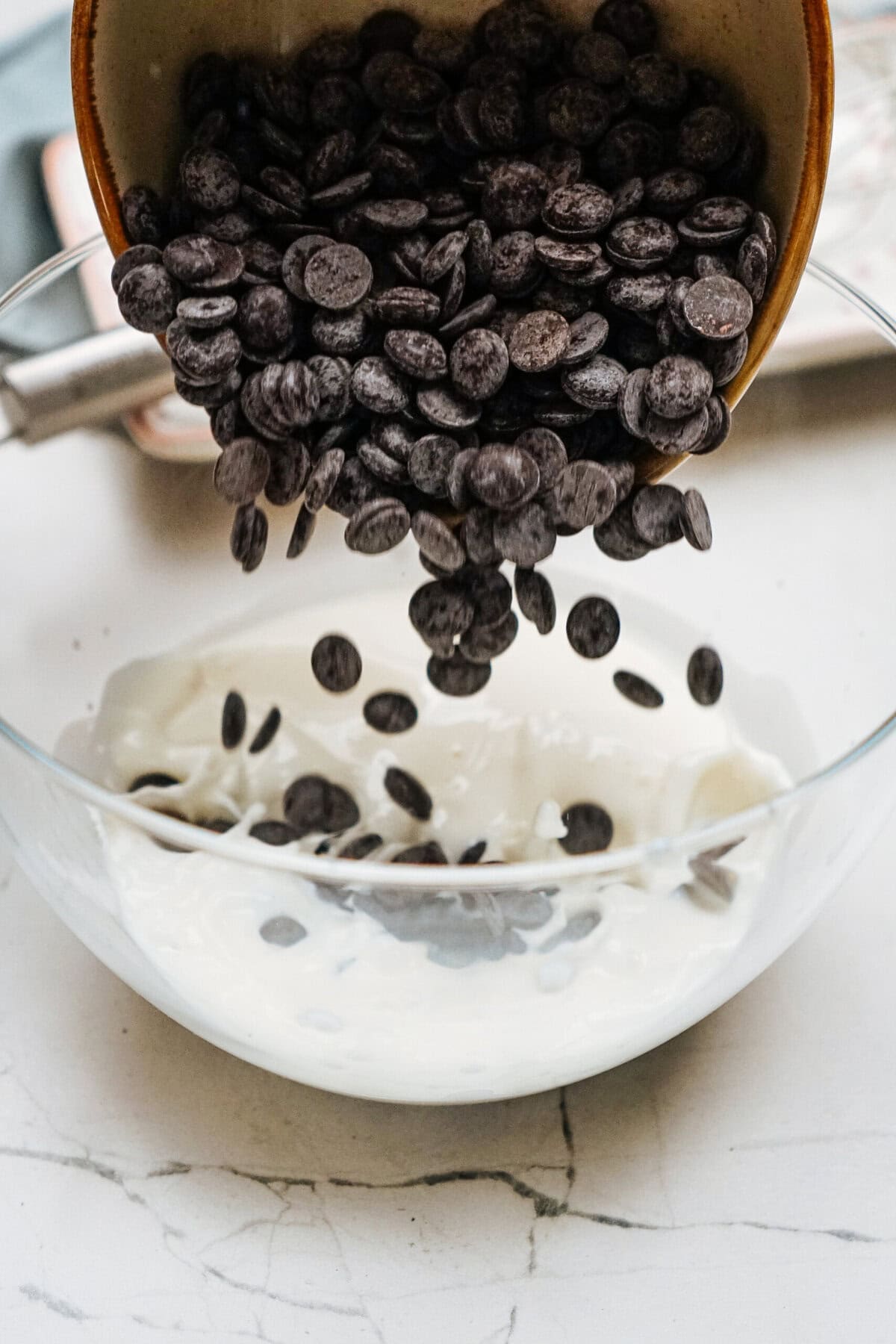 Chocolate chips being poured into a bowl of white cream on a marble surface.