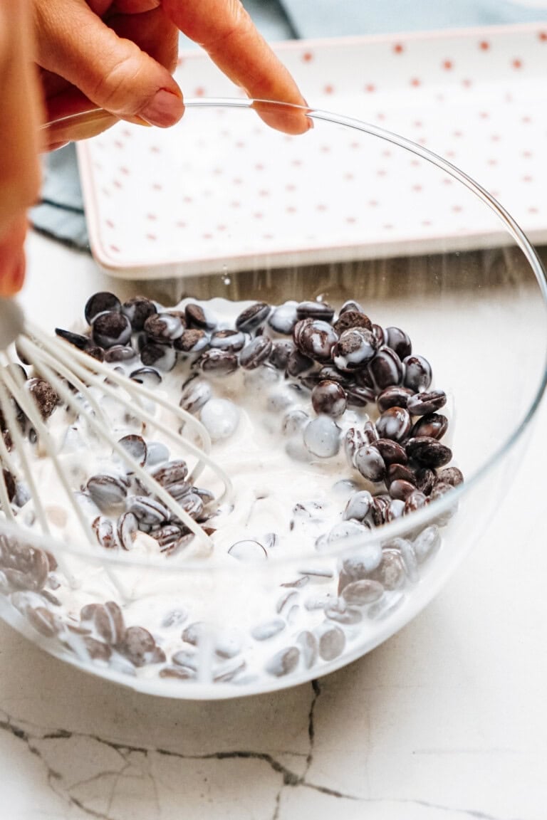 A person whisks dark chocolate chips into a bowl of thick cream on a marble countertop.