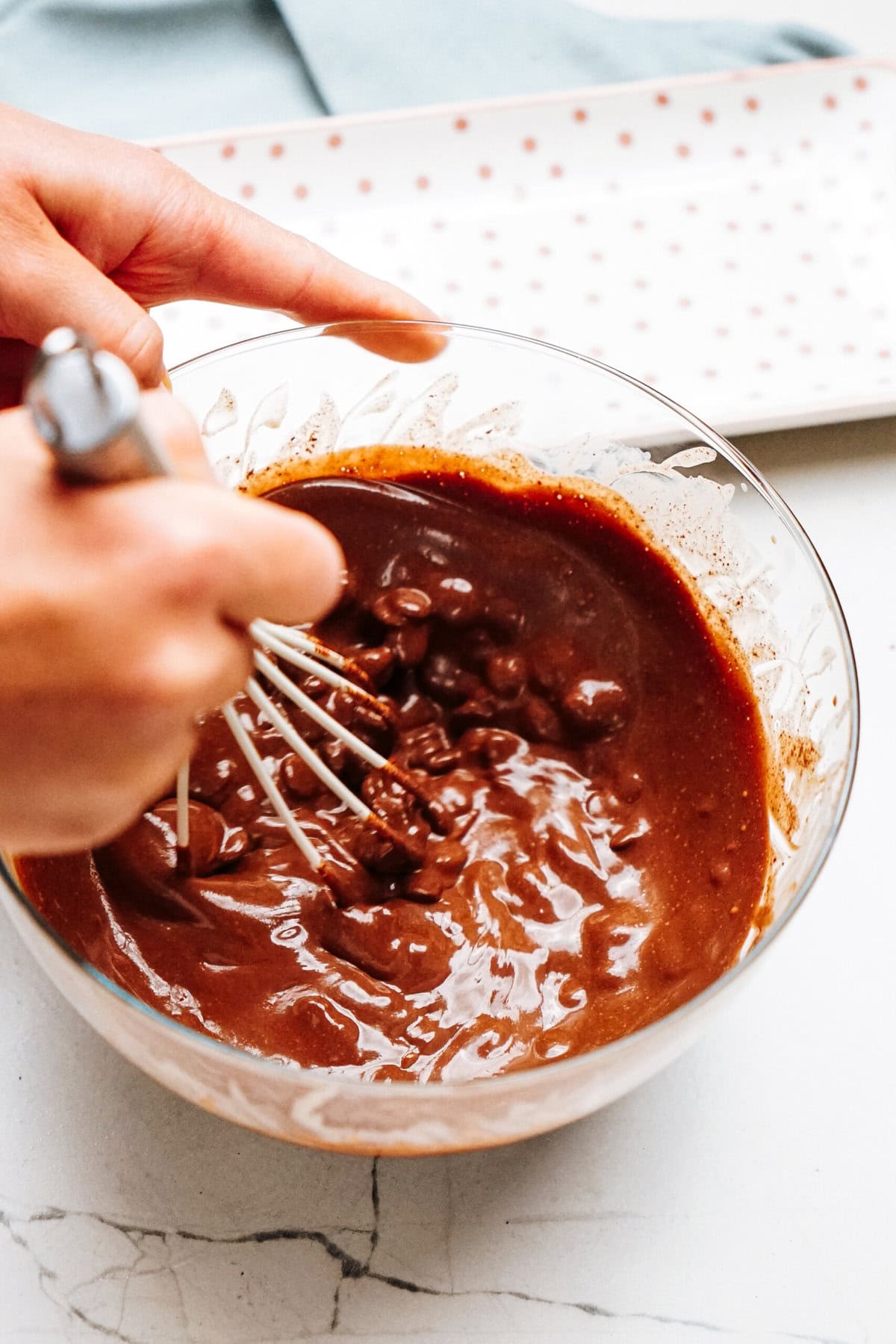 A person whisks a mixture of chocolate batter and chocolate chips in a glass bowl on a marble surface.