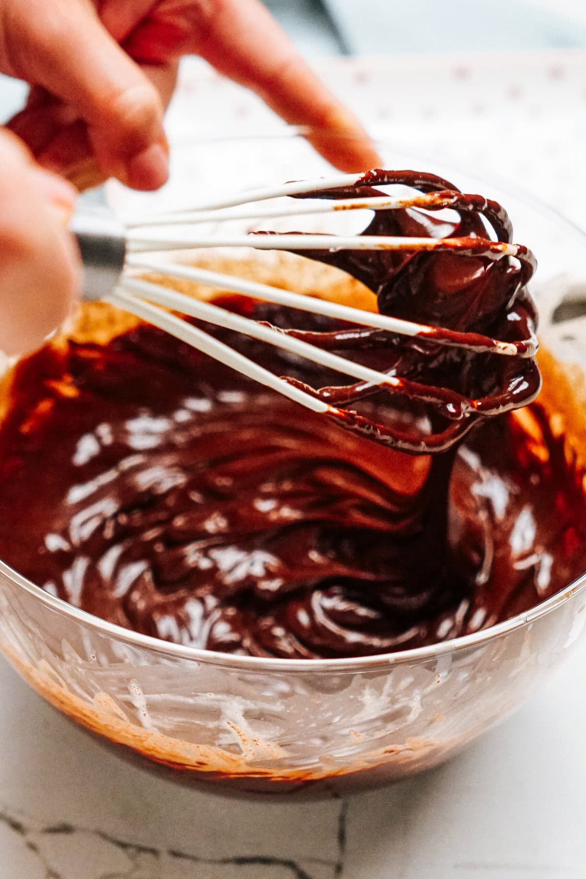 A person mixing chocolate batter in a glass bowl with a whisk, on a white countertop.