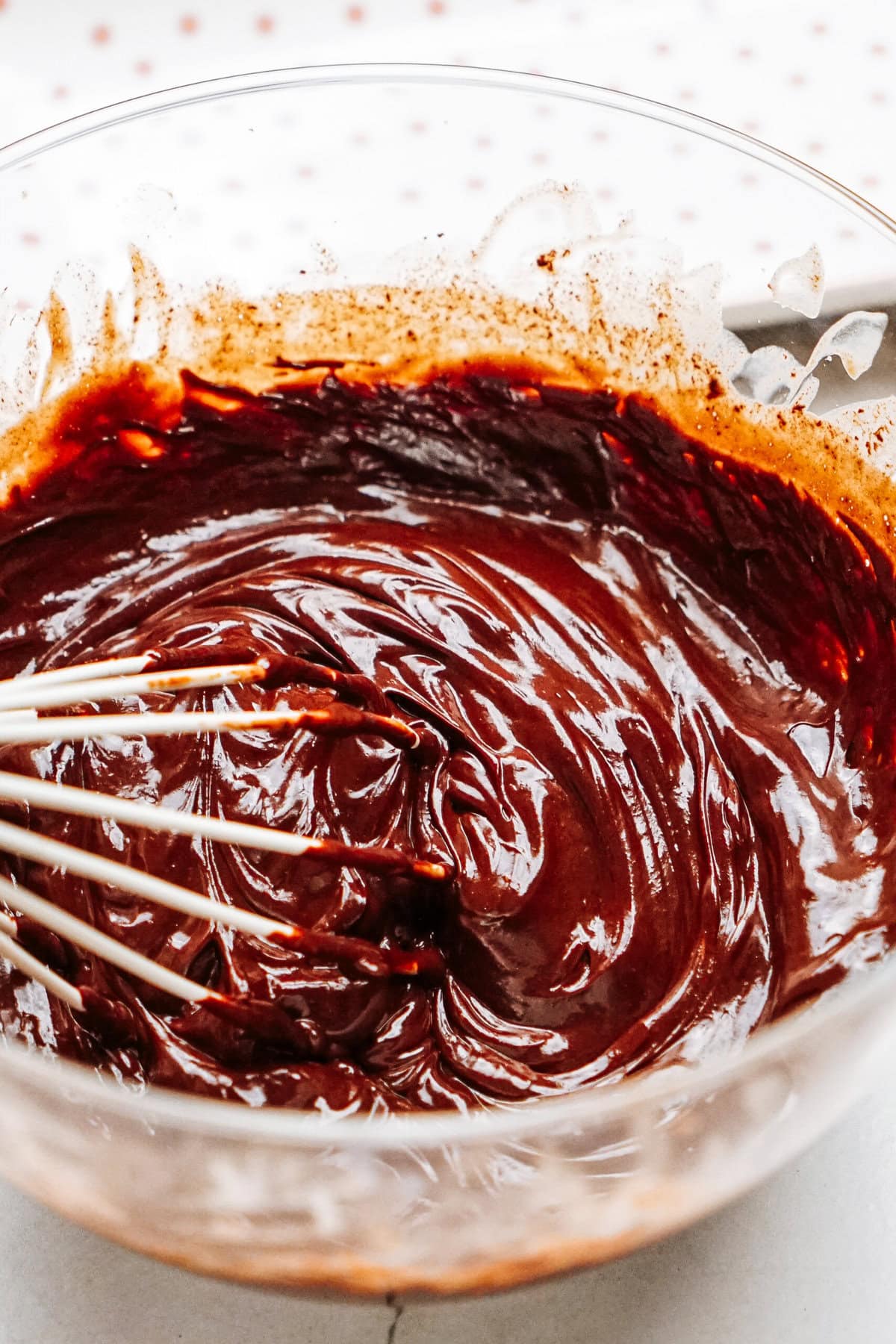 Close-up of chocolate batter being mixed in a glass bowl with a whisk, against a light background.