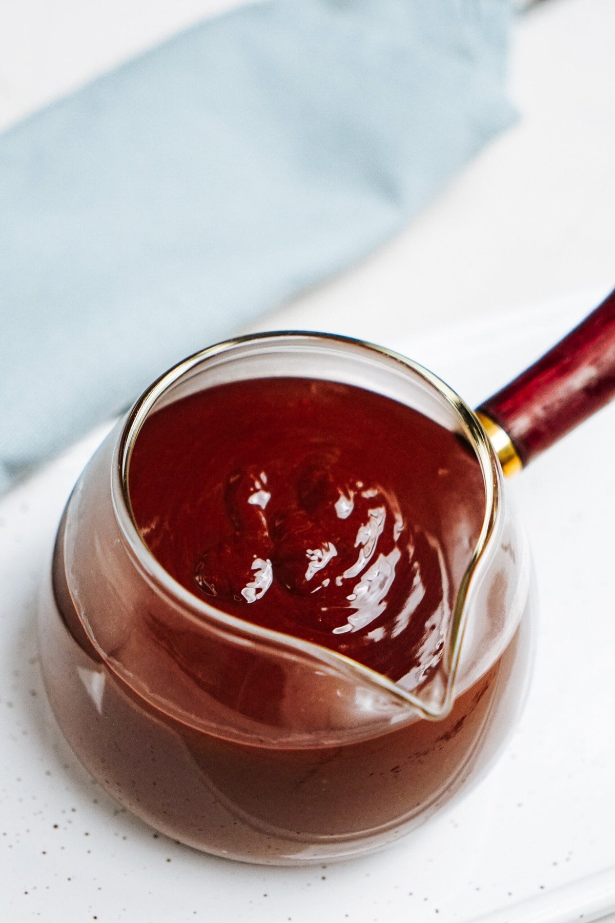 Glass pitcher filled with thick, chocolate ganache sauce on a white speckled surface.