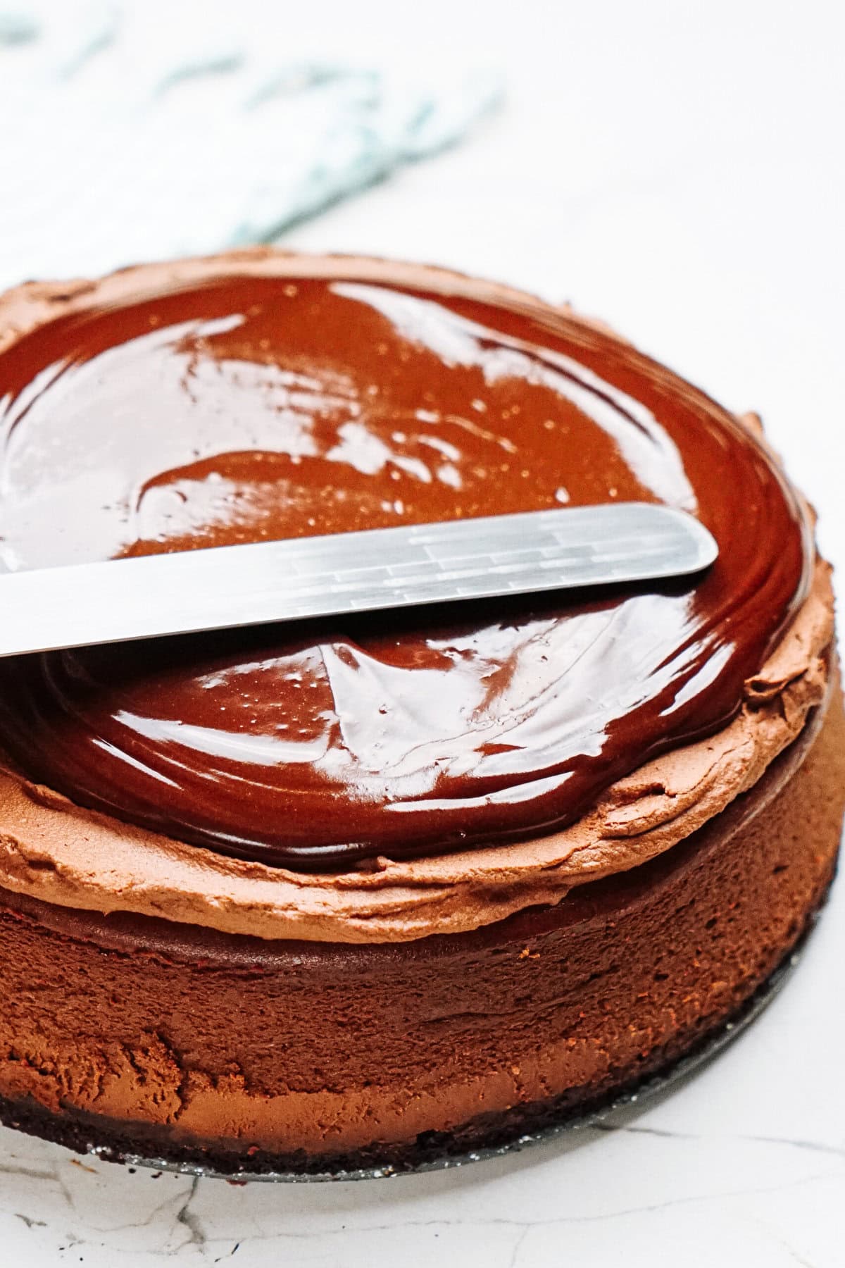 A chocolate cake is being frosted with ganache using a metal spatula on a marble surface.