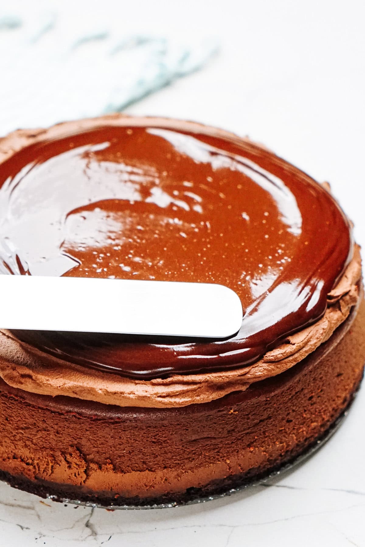 A spatula gracefully spreads rich chocolate ganache over the top of a round cheesecake resting on a pristine white surface.