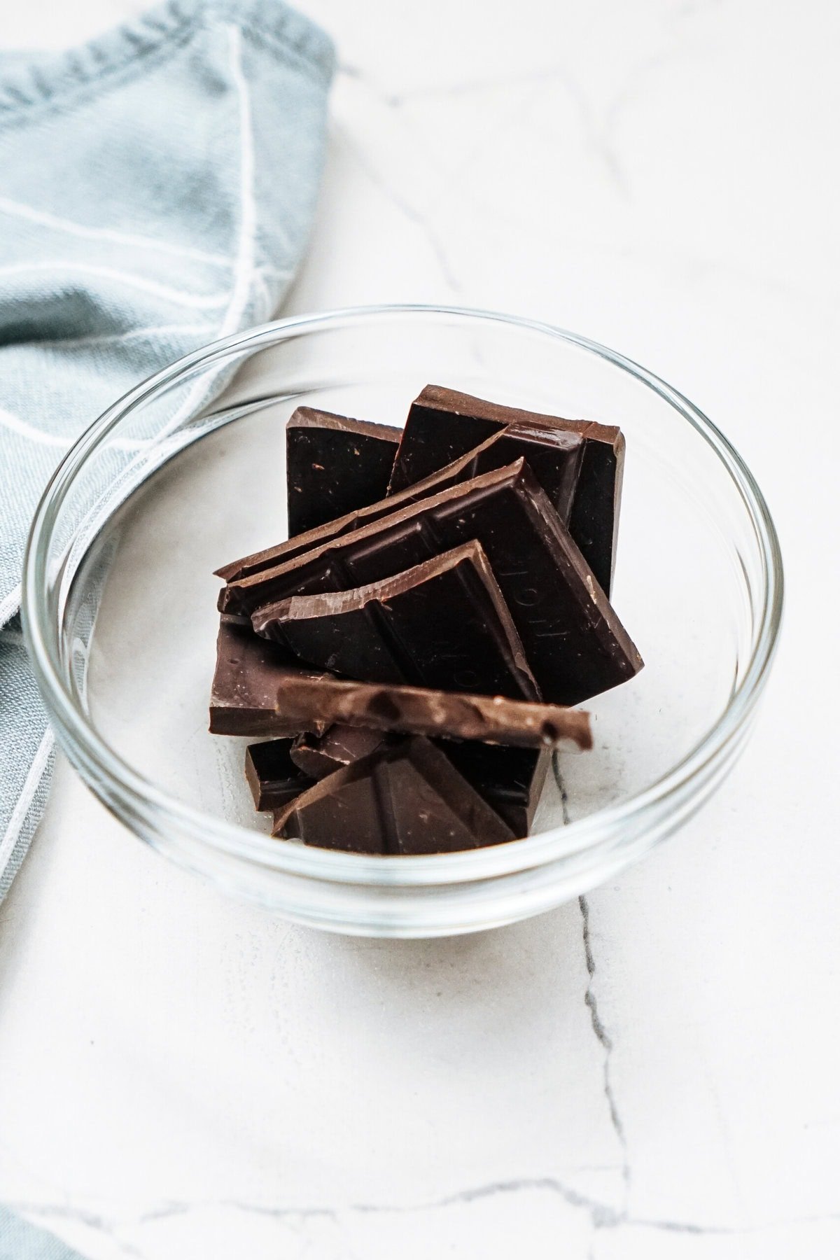 A glass bowl filled with roughly broken pieces of dark chocolate on a white marble surface. A corner of a light blue cloth is visible next to the bowl.