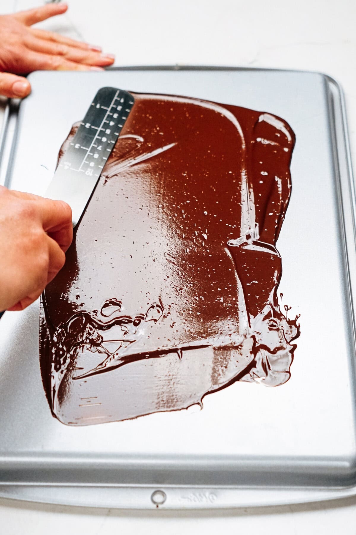 Hands using a spatula to spread melted chocolate on a metal baking sheet.