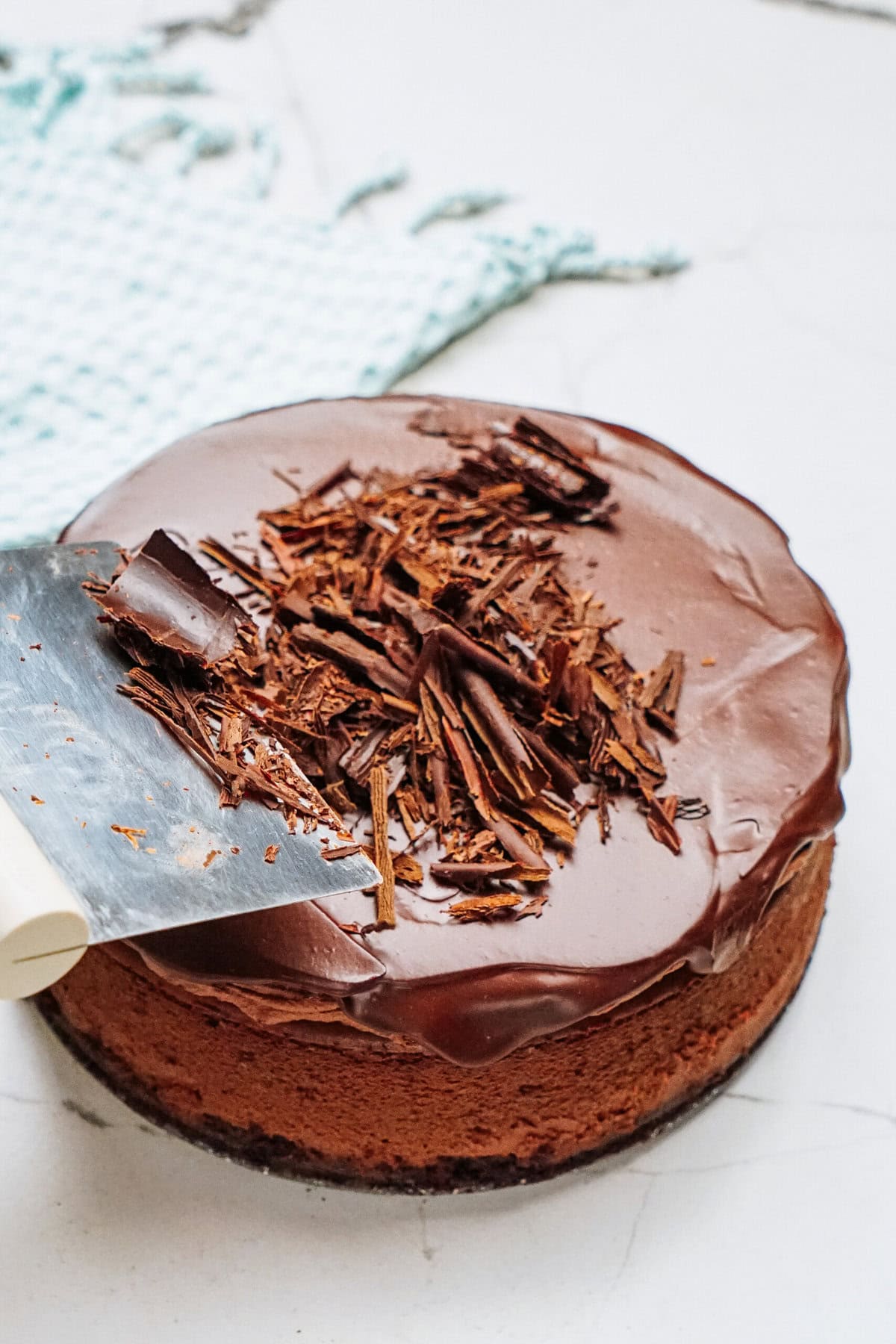 Chocolate cake topped with chocolate shavings, partially sliced on a white surface with a light blue cloth in the background.