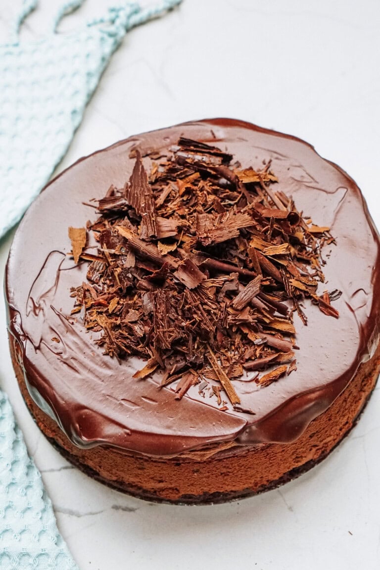 A round chocolate cake topped with chocolate shavings, placed on a white surface near a light blue textured cloth.