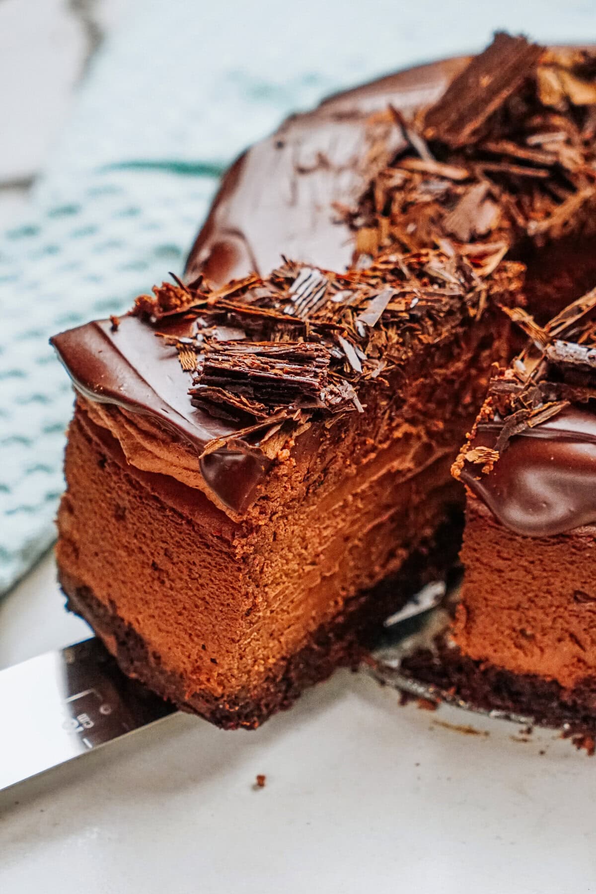 A slice of chocolate cake with creamy layers and a chocolate shavings topping is served on a cake server.