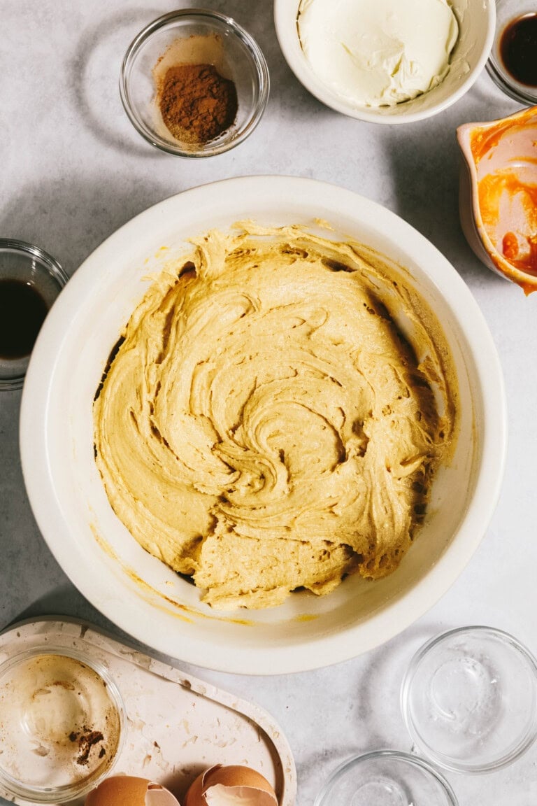 A mixing bowl filled with a creamy yellow batter, surrounded by ingredients including eggshells, spices, vanilla extract, and butter on a countertop.