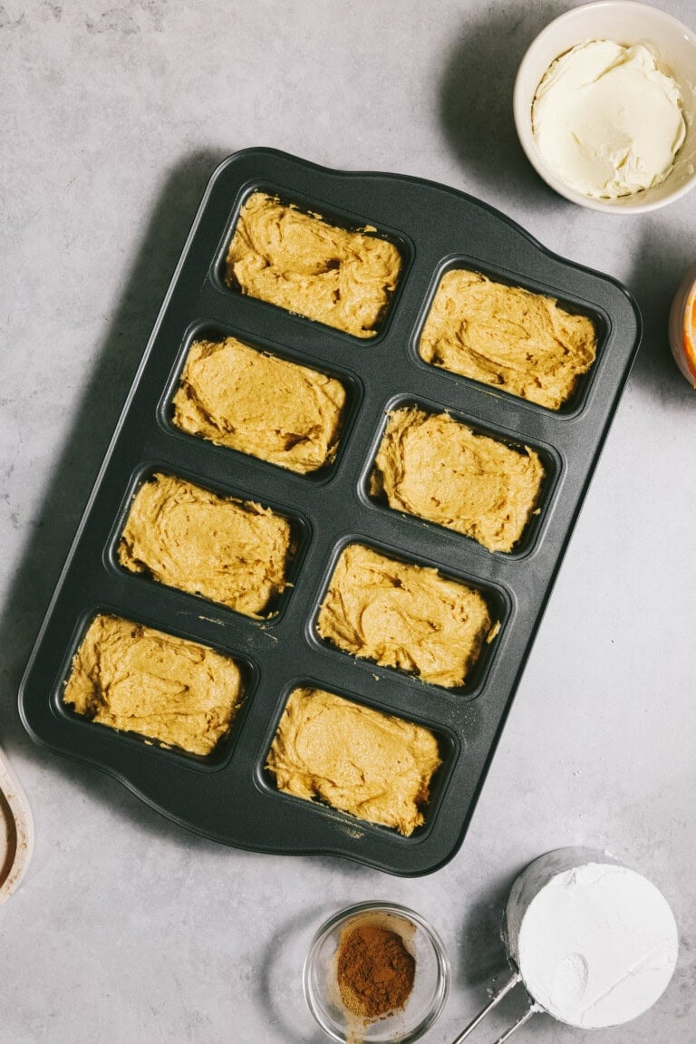 A baking tray with eight rectangular molds filled with pumpkin batter. Surrounding the tray are various baking ingredients, including flour, ground cinnamon, and a bowl of cream cheese mixture.