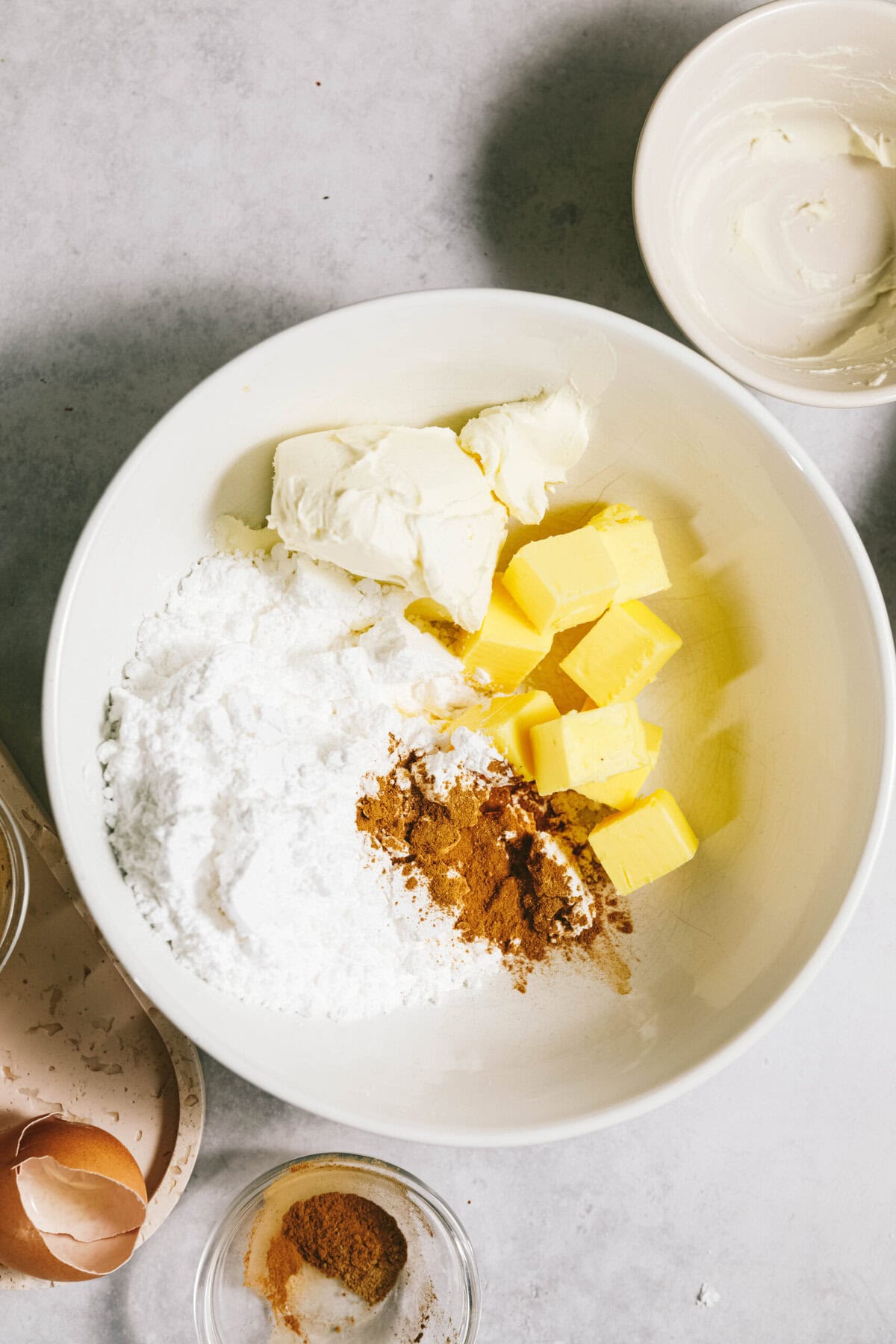 A mixing bowl contains butter, cream cheese, powdered sugar, and spices. Surrounding are a cracked egg, a small bowl of spices, and a bowl with cream cheese remnants.