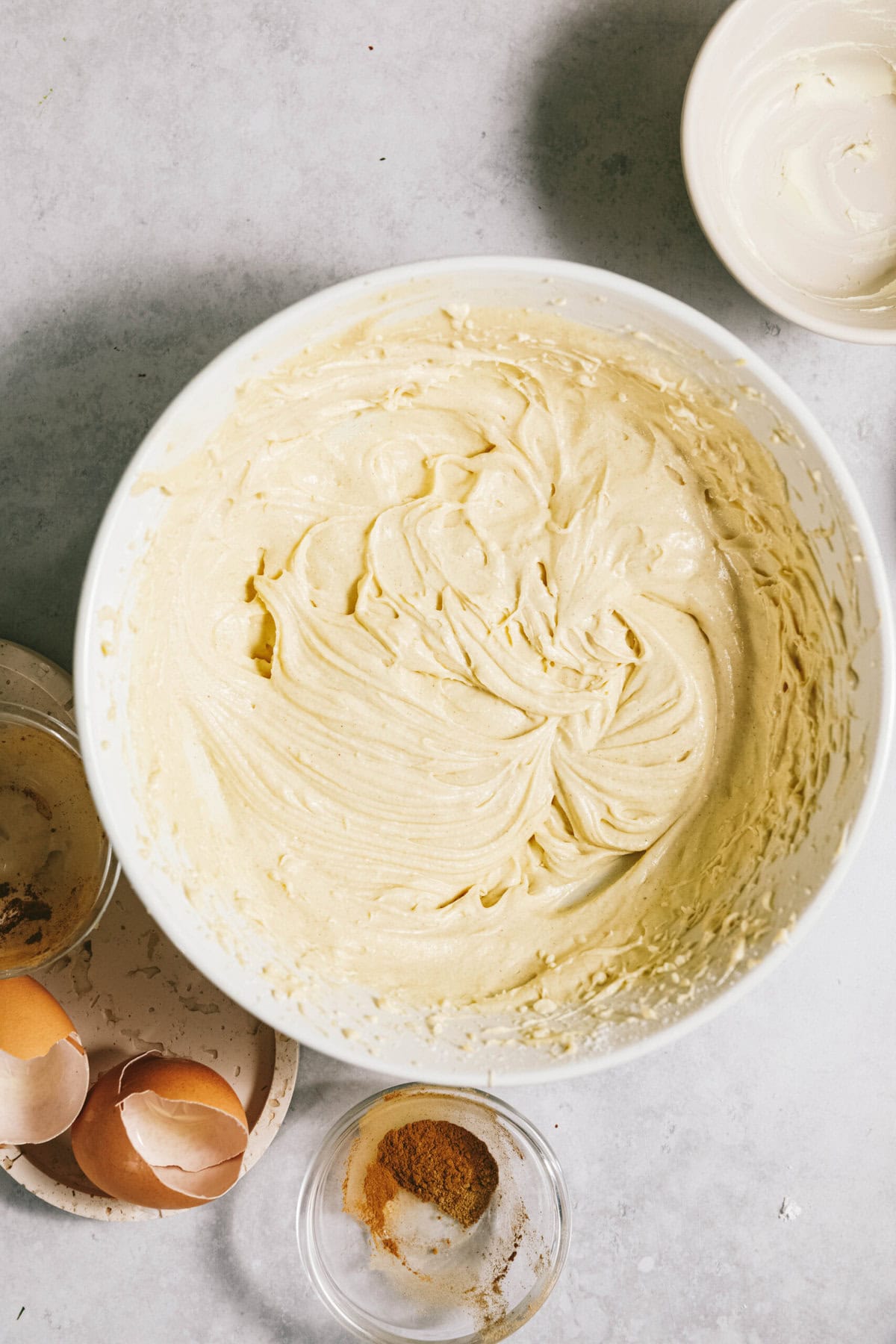 A bowl full of dough, along with broken eggshells, a small bowl with a spice, and an empty bowl on a light countertop.