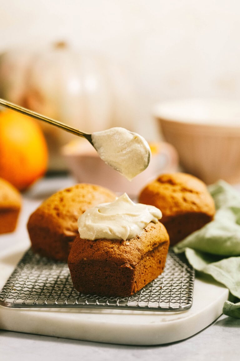 A spoon spreading frosting on mini loaves of bread on a cooling rack, with pumpkins and a green cloth in the background.