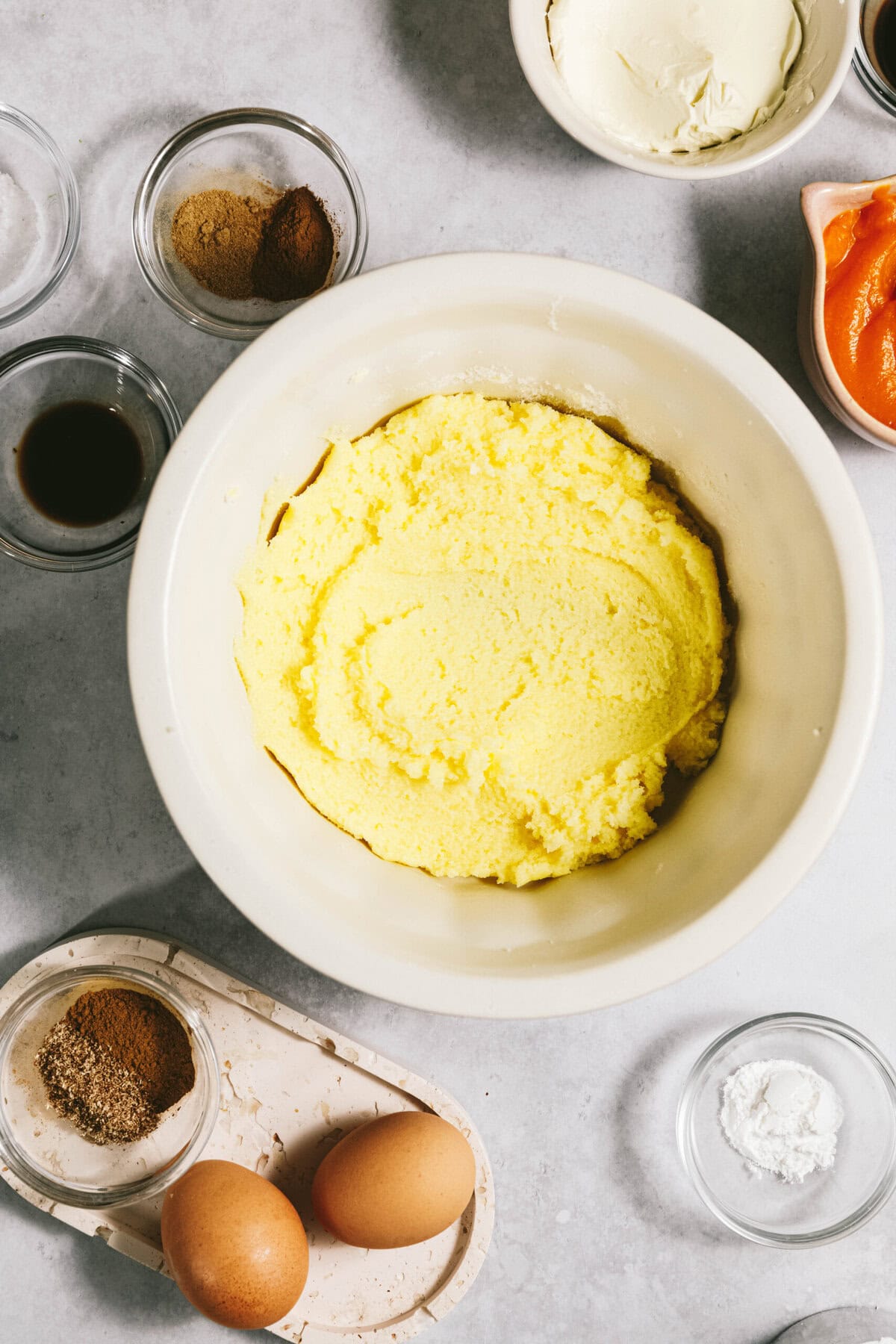 A bowl of yellow mixture is surrounded by various baking ingredients, including eggs, spices, vanilla extract, and pumpkin puree on a countertop.