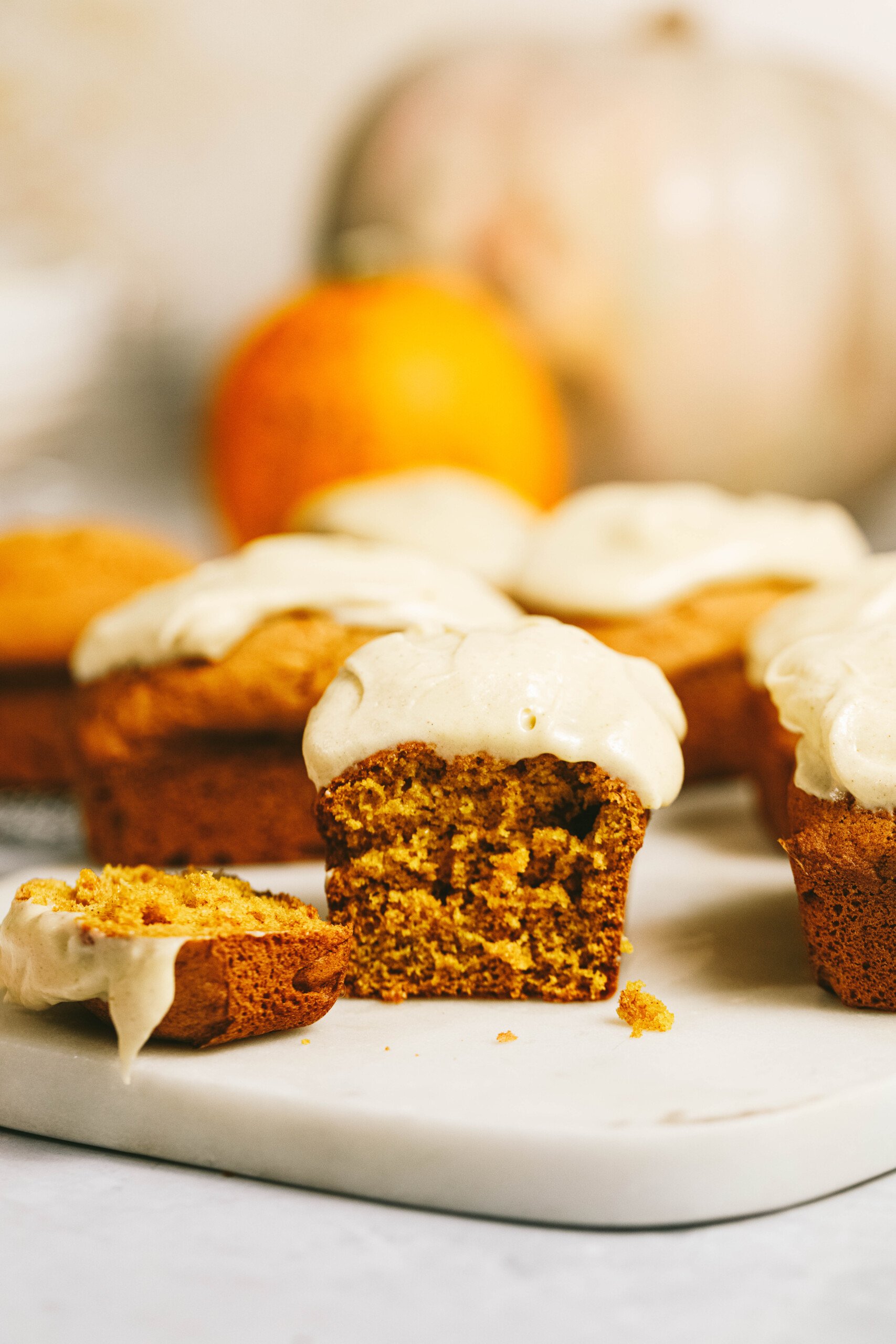 Close-up of several pumpkin muffins covered in white icing, one partially eaten, on a white surface with a pumpkin and orange fruit in the background.