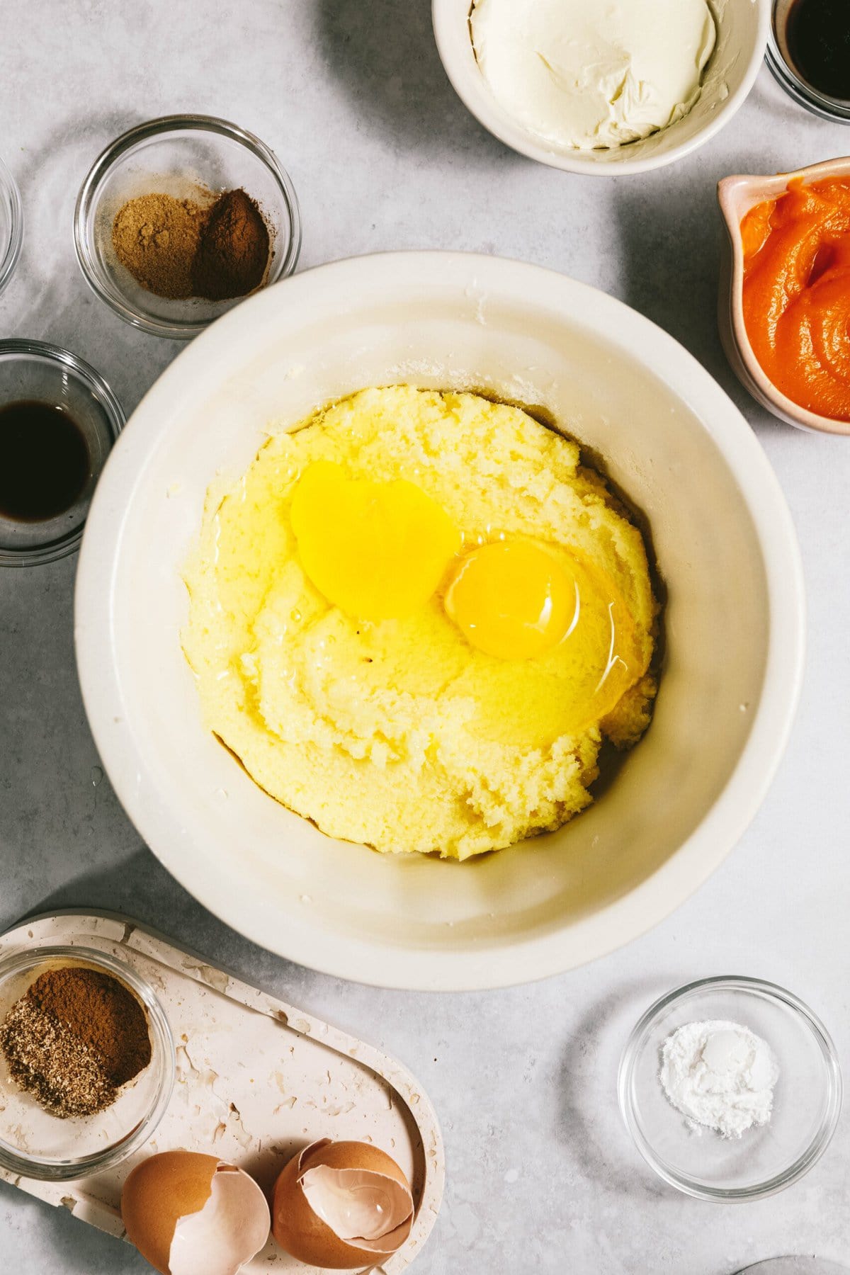 A mixing bowl with a batter and two cracked eggs on top, surrounded by small bowls containing various ingredients, including spices, vanilla, pumpkin puree, and empty eggshells.