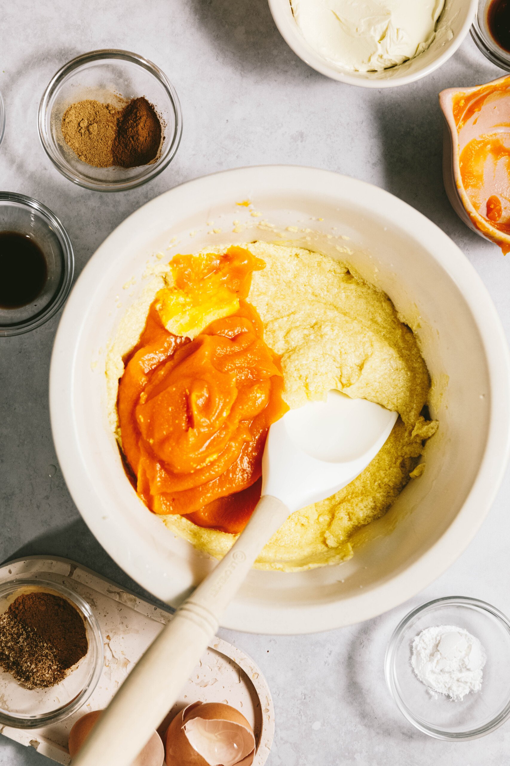 Mixing bowl with pumpkin puree and batter, surrounded by small bowls containing spices, vanilla extract, and egg shells.