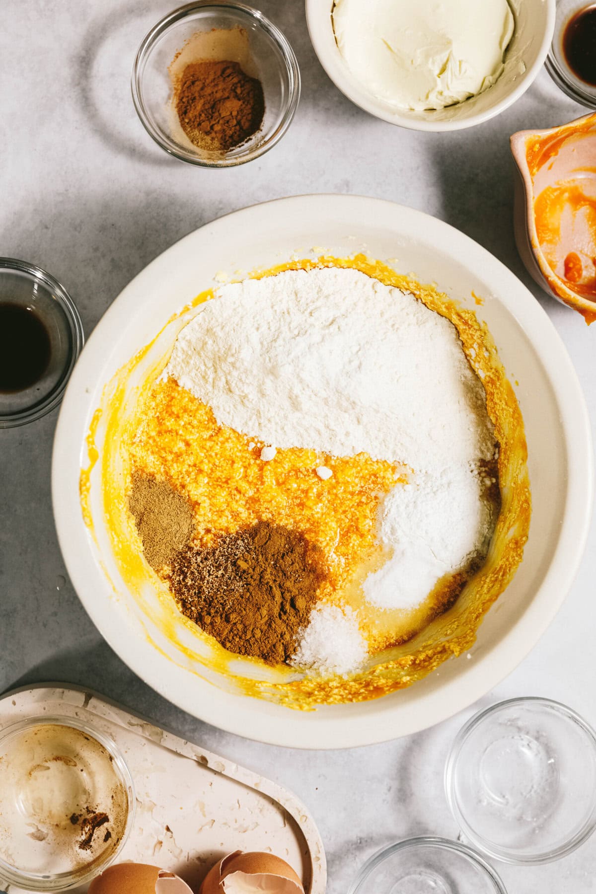 Mixing bowl with flour, spices, and wet ingredients on a countertop, surrounded by empty bowls and broken eggshells.