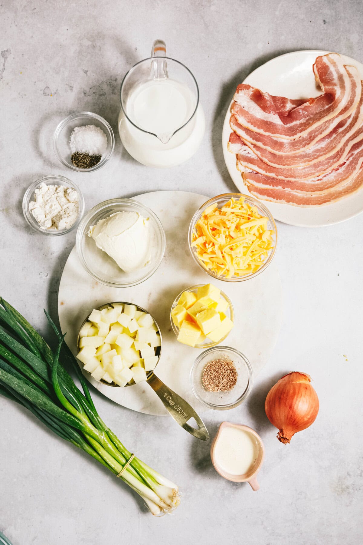 Ingredients on a table include green onions, diced potatoes, butter, grated cheese, sour cream, bacon, milk, flour, salt, pepper, and an onion.