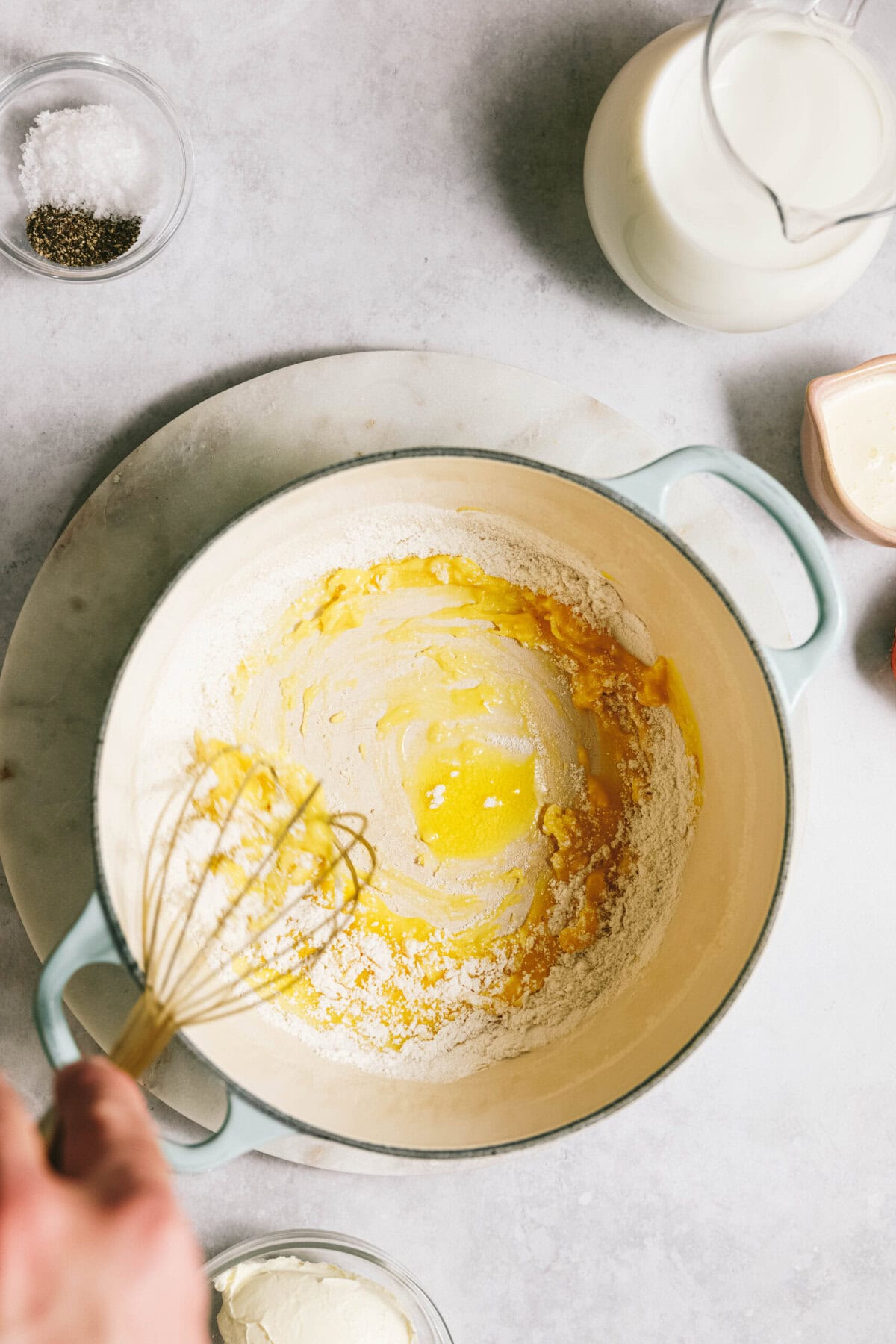 A hand whisking melted butter into flour in a light blue pot, surrounded by bowls of salt, pepper, milk, and other ingredients on a light surface.