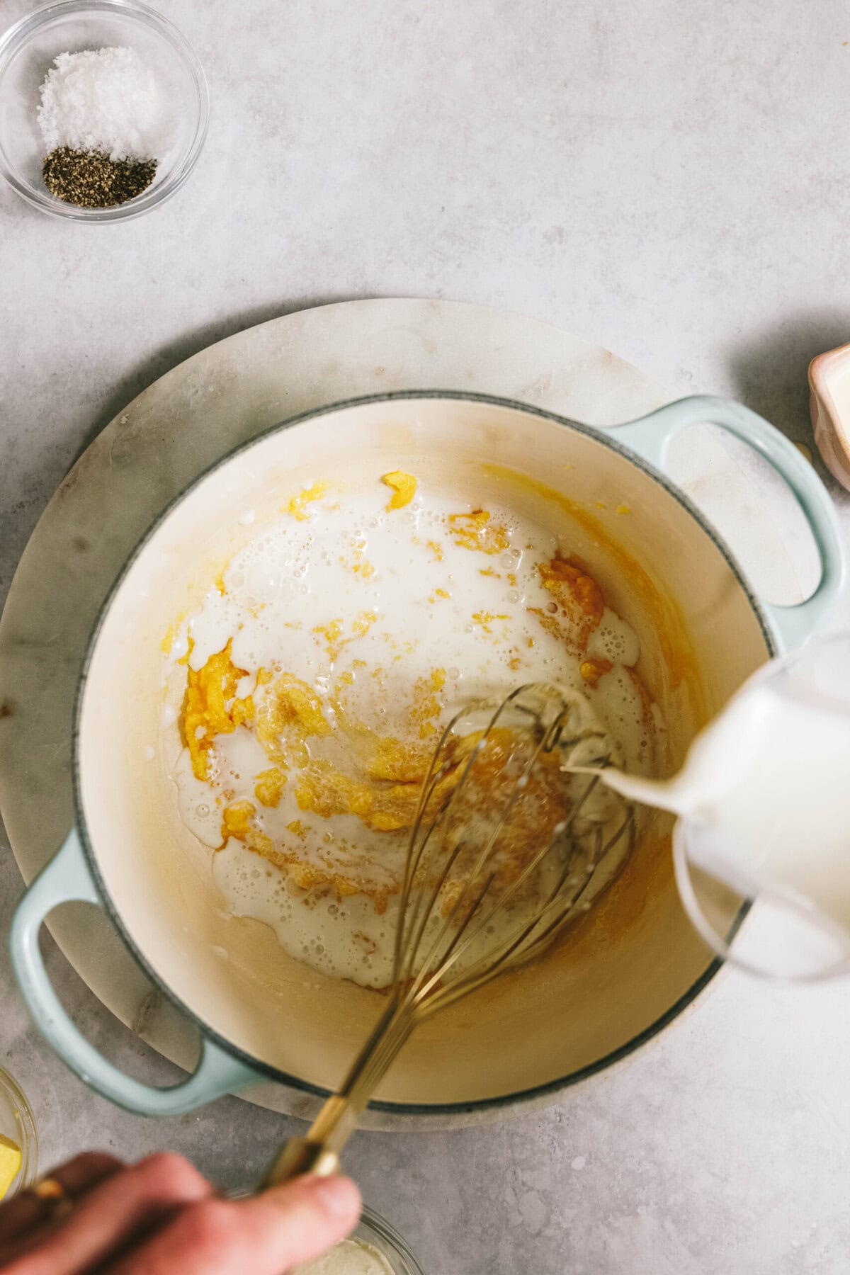 Whisk stirring milk into a buttery flour mixture in a large pot, with salt and pepper bowls nearby on a marble surface, preparing the base for a delicious potato casserole.