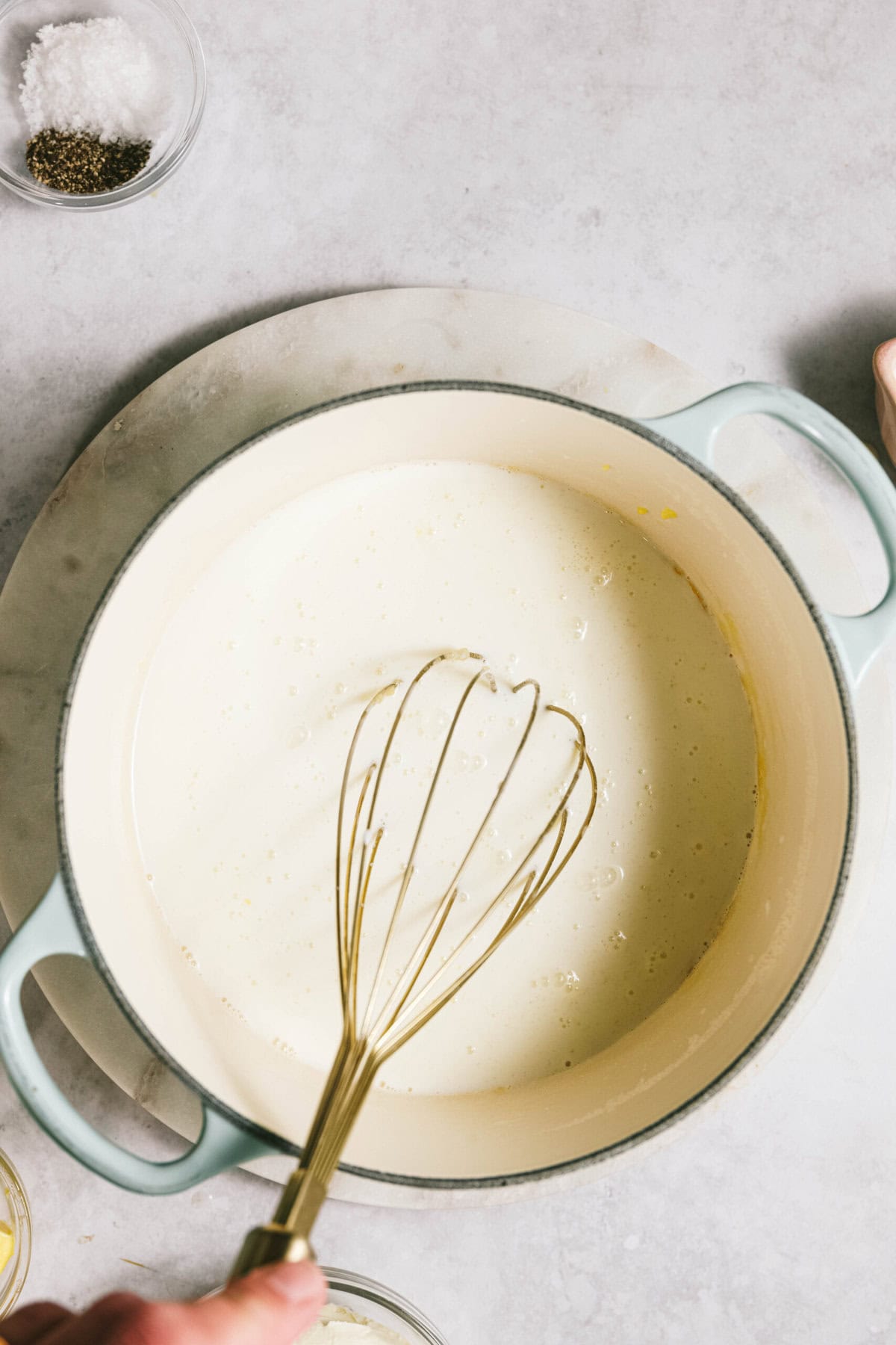 A whisk stirs creamy sauce in a light blue pot, the perfect base for a savory potato casserole. Nearby, small bowls with salt and pepper rest on the marble surface.