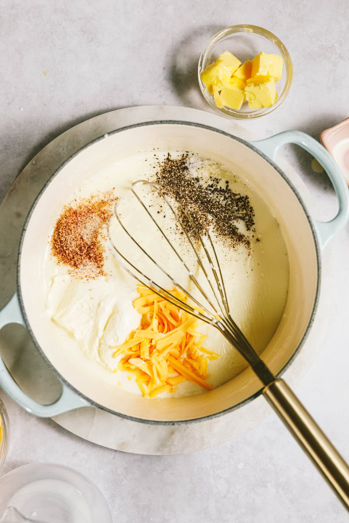 A pot with creamy sauce, grated cheese, ground pepper, and nutmeg being whisked. Butter in small bowl nearby on a light surface.