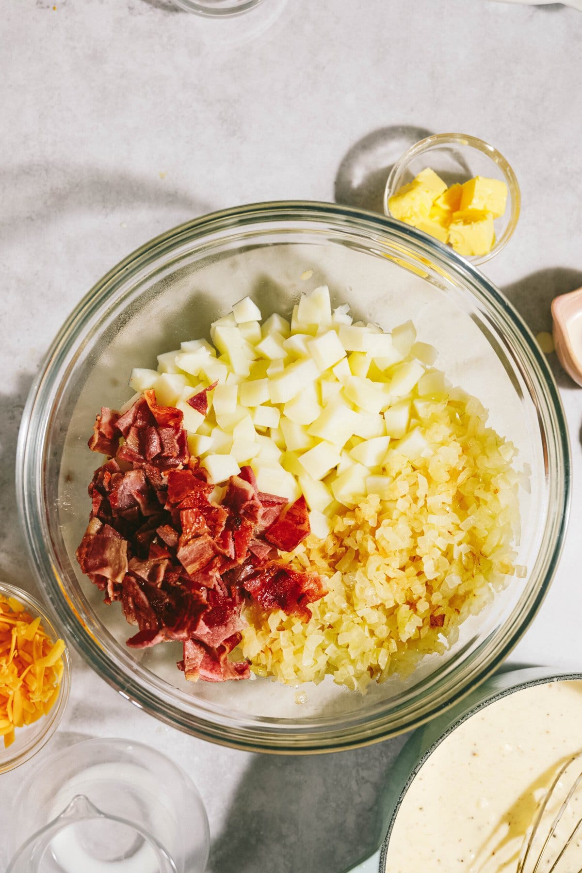 A glass bowl with diced potatoes, chopped bacon, and onions. Surrounding ingredients include bowls of butter, cheddar cheese, and a cream mixture on a light countertop.