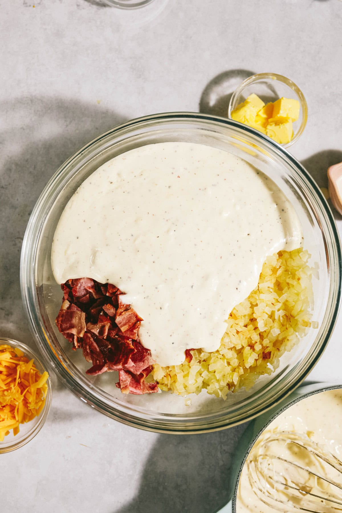 A bowl with chopped bacon, onions, and creamy sauce. Nearby are small bowls of grated cheese and butter.