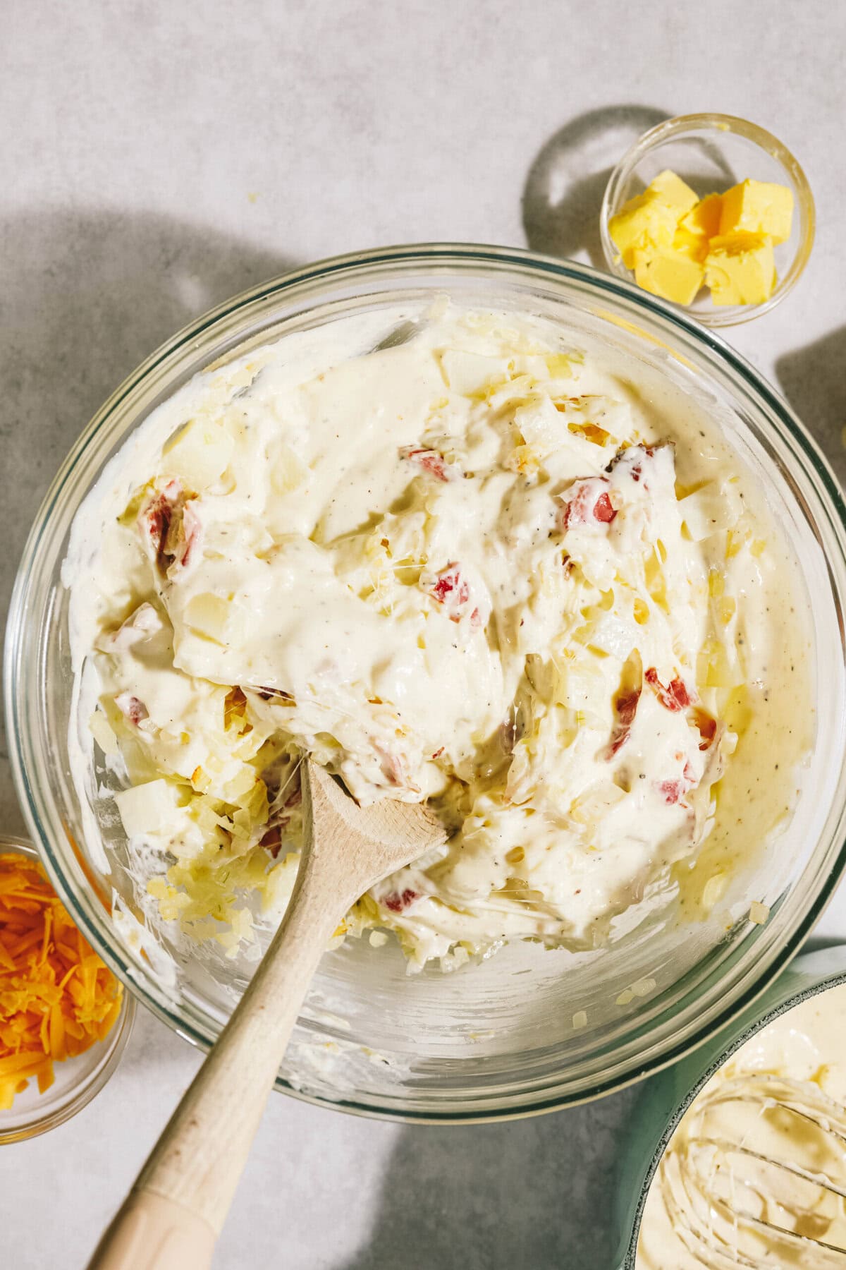 Bowl of creamy mixture with shredded cheese and chopped bacon, wooden spoon inside; small bowls of butter and more cheese nearby.