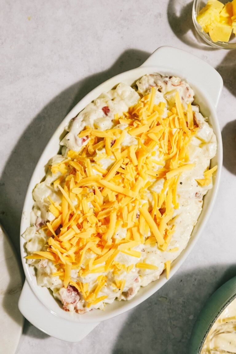 A casserole dish filled with creamy potato salad topped with shredded cheddar cheese, placed on a light-colored surface.