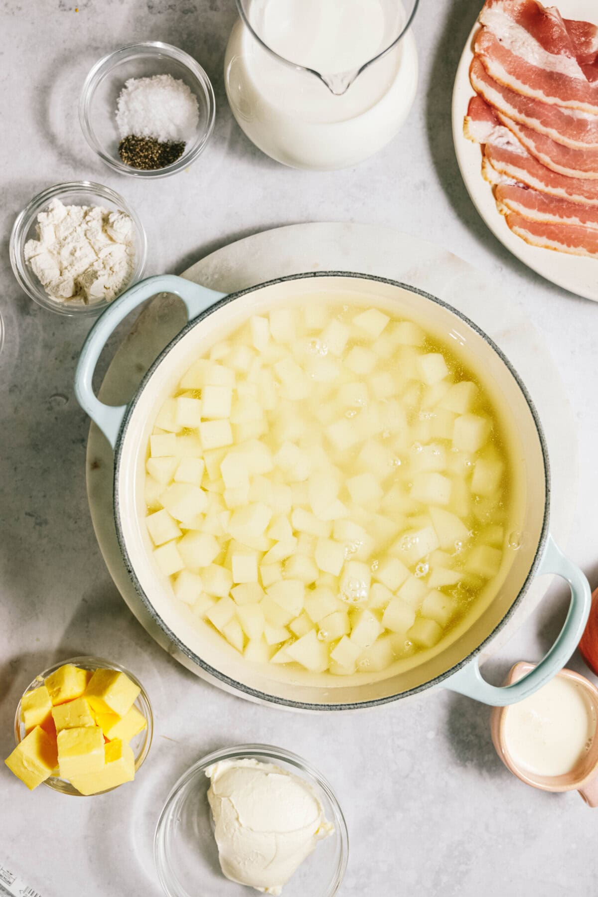 Cooking ingredients on a countertop: diced potatoes in a pot, bacon on a plate, cream in a jug, butter, flour, pepper, and salt in bowls, with a small bowl of sour cream.