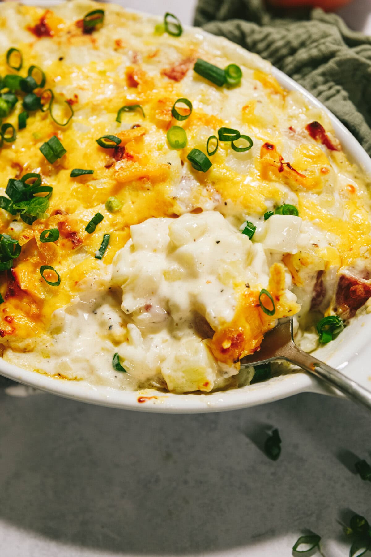 A dish of baked potato casserole topped with melted cheese, chopped green onions, and a serving spoon inserted.