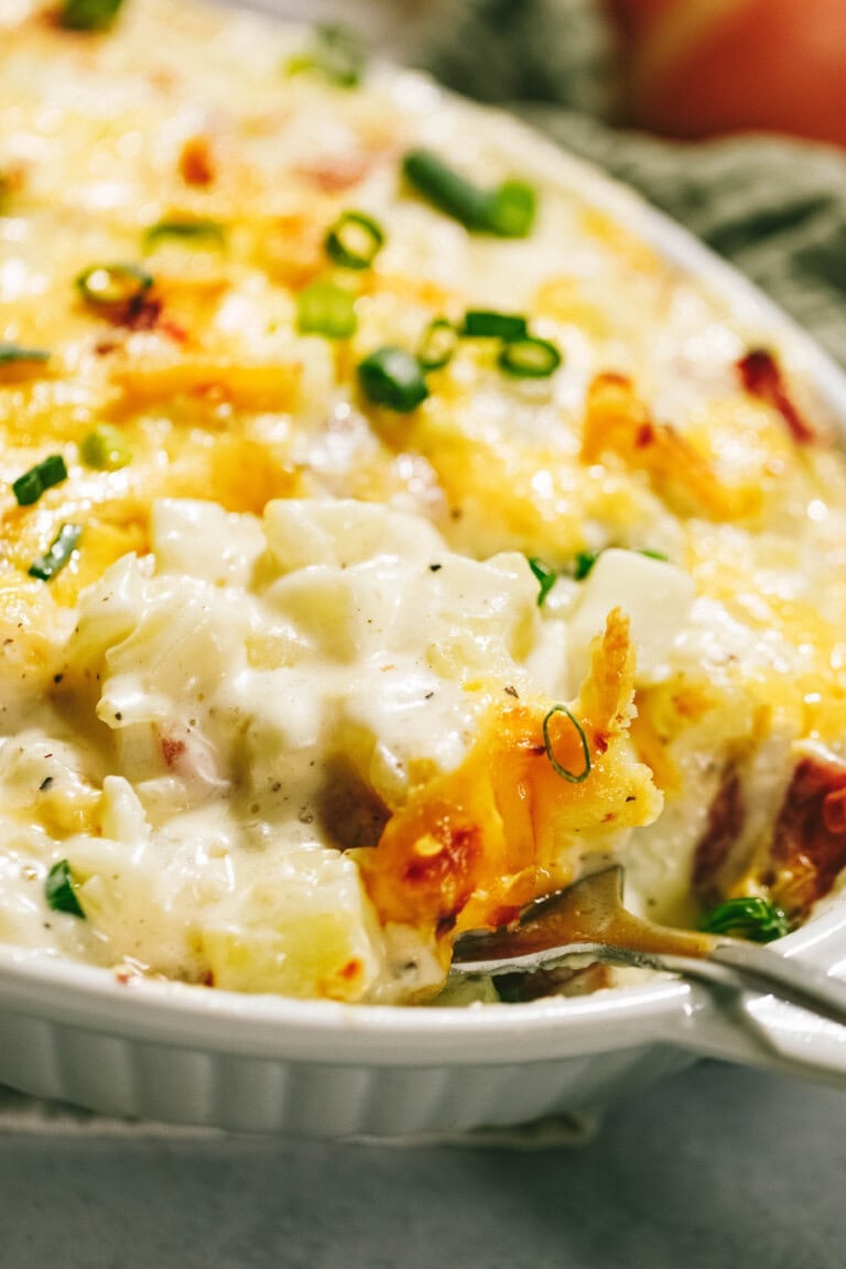 Close-up of a cheesy potato casserole with a creamy texture and garnished with chopped green onions. A spoon is partially dipped into the dish.