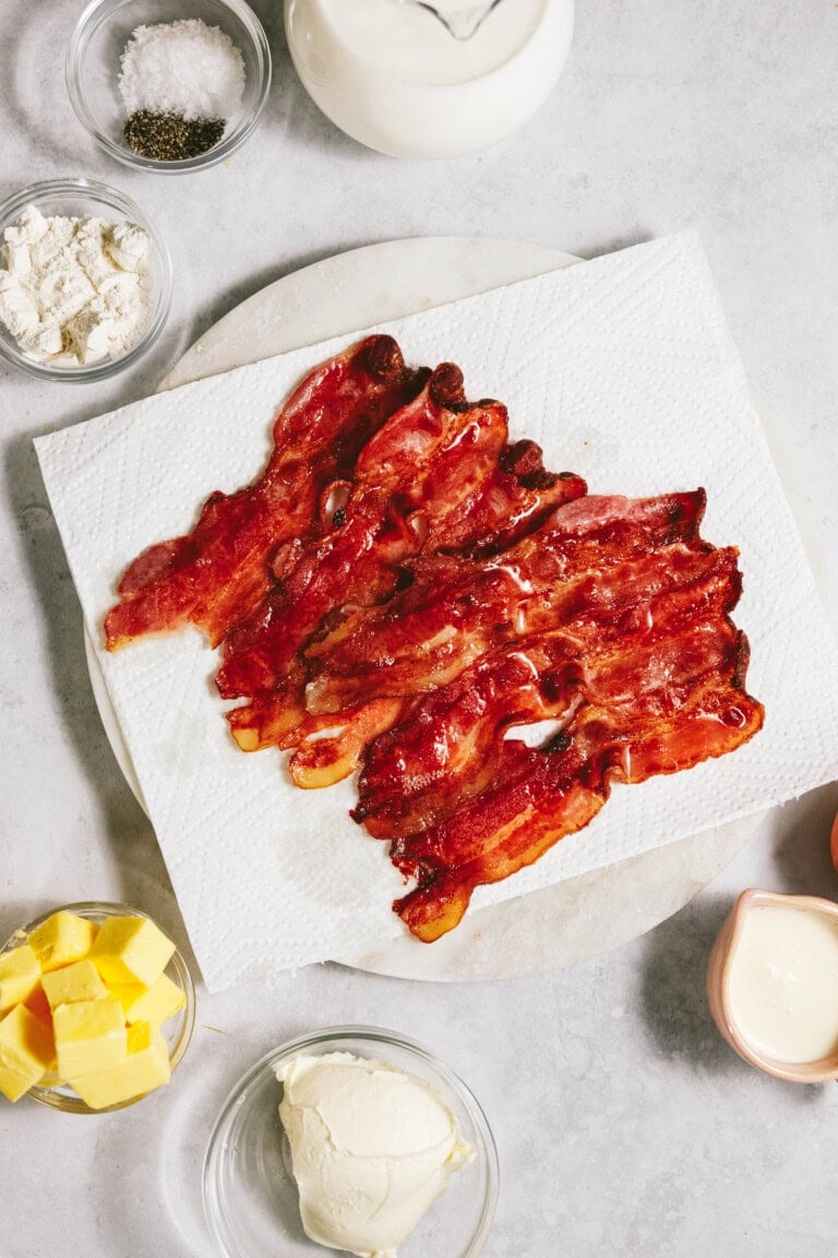 Crispy bacon strips on a paper towel surrounded by small bowls of ingredients like butter, cream, and seasonings on a light countertop.