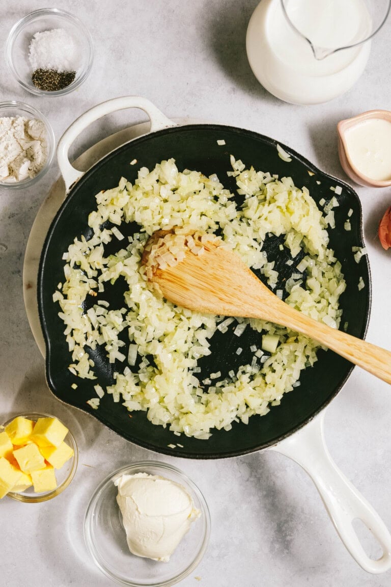 Chopped onions frying in a white skillet with a wooden spoon. Surrounding the skillet are bowls of butter, cream, flour, and seasonings.