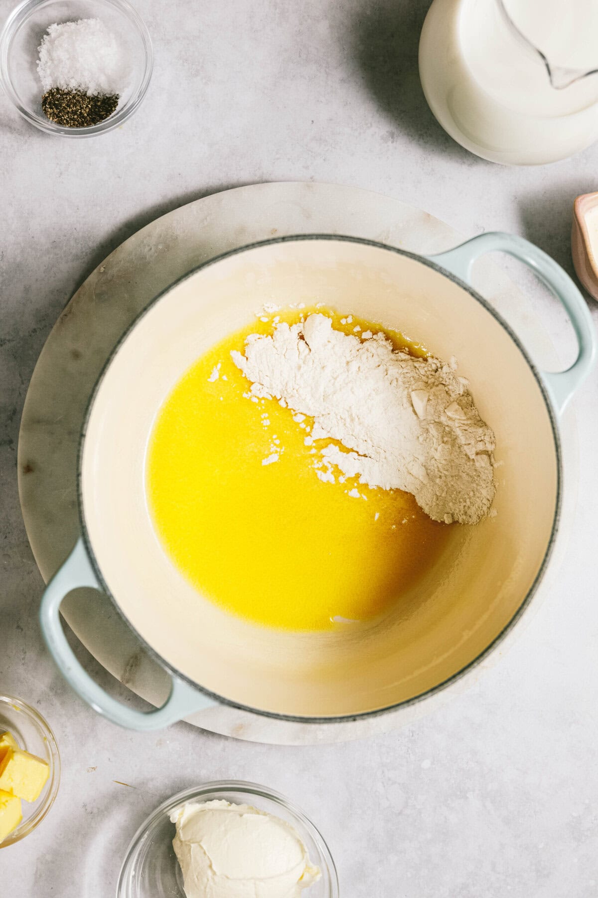 A pot on a marble surface containing melted butter and flour, surrounded by small bowls of salt, pepper, cream, and butter.