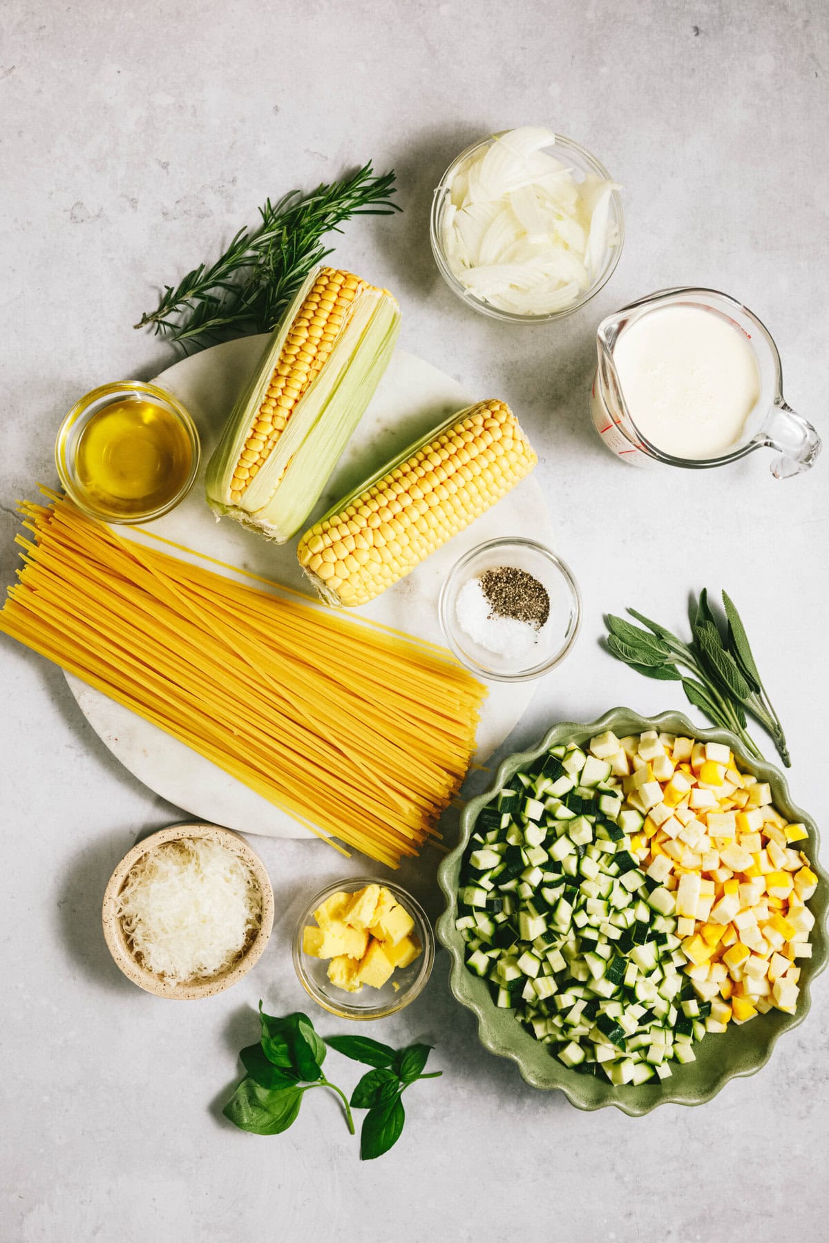 On the countertop, ingredients for a summer squash pasta await: uncooked spaghetti, corn, chopped zucchini and summer squash, butter, onion, cream, rosemary, sage, olive oil, salt, pepper, grated cheese, and basil.