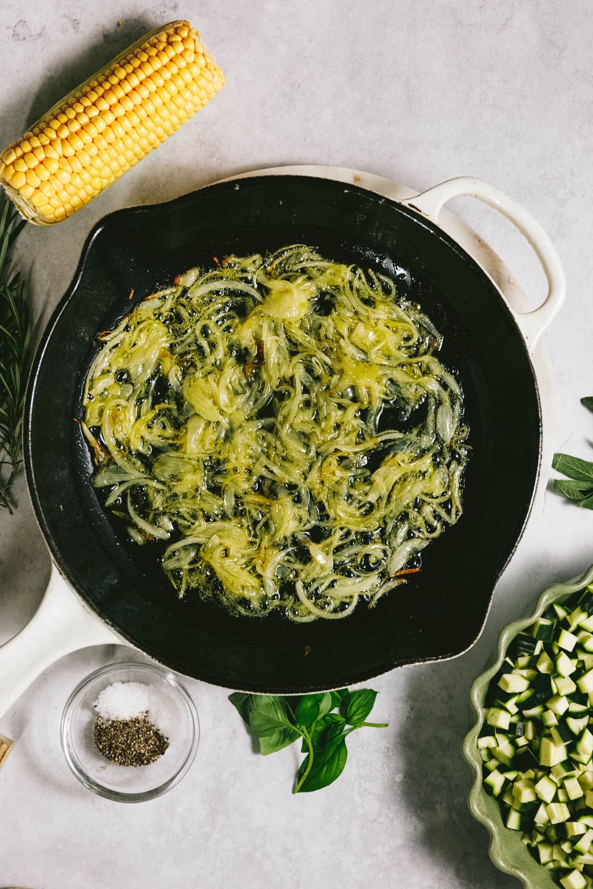 Onions sautéing in a pan with corn beside a bowl of chopped zucchini and a dish of salt and pepper, all waiting to become the perfect summer squash pasta.