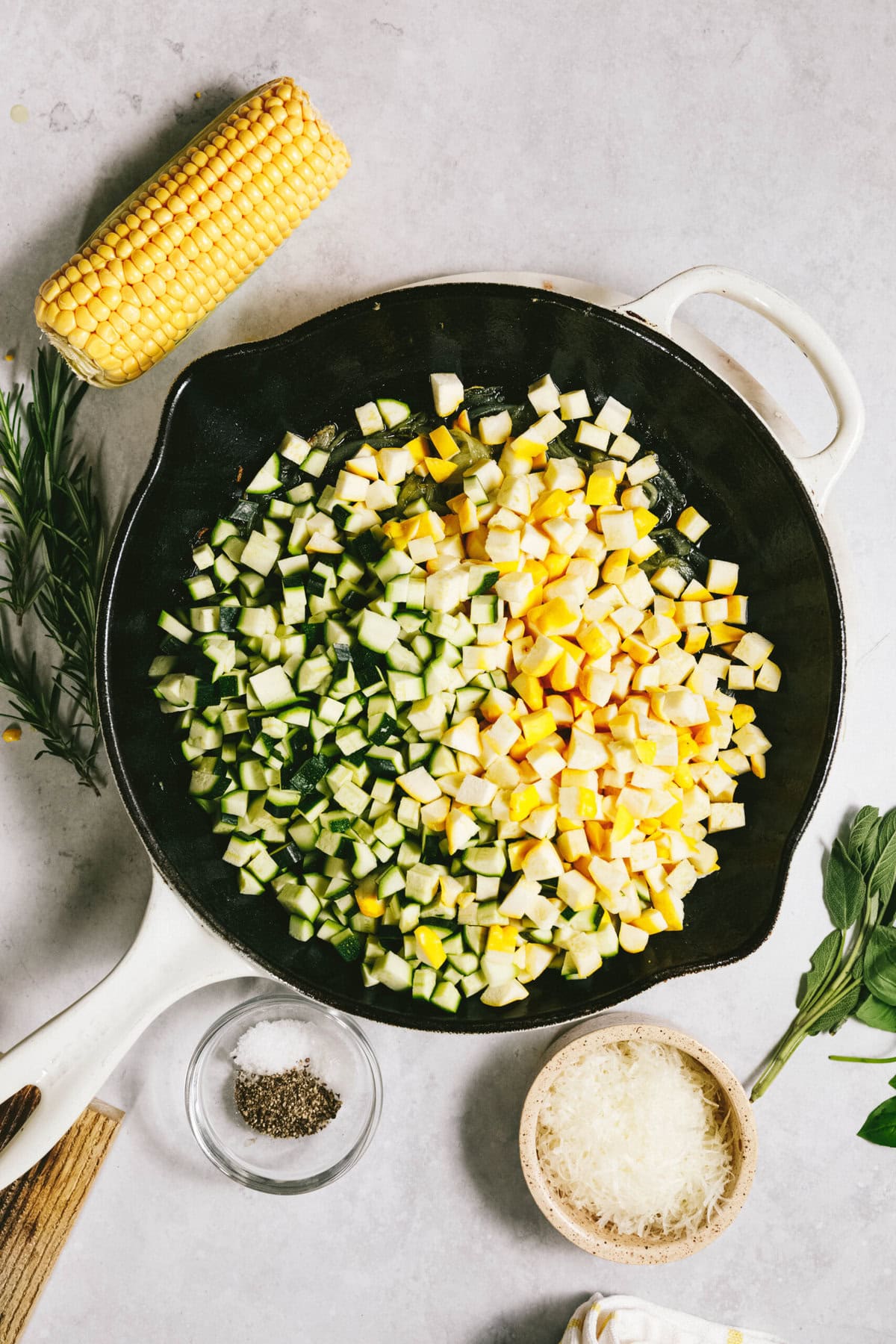 Chopped zucchini and squash mingle in a pan, alongside corn, rosemary, sage, and a small bowl of cheese. A bowl of salt and pepper completes the scene on a light countertop, perfect for crafting a breezy summer squash pasta dish.