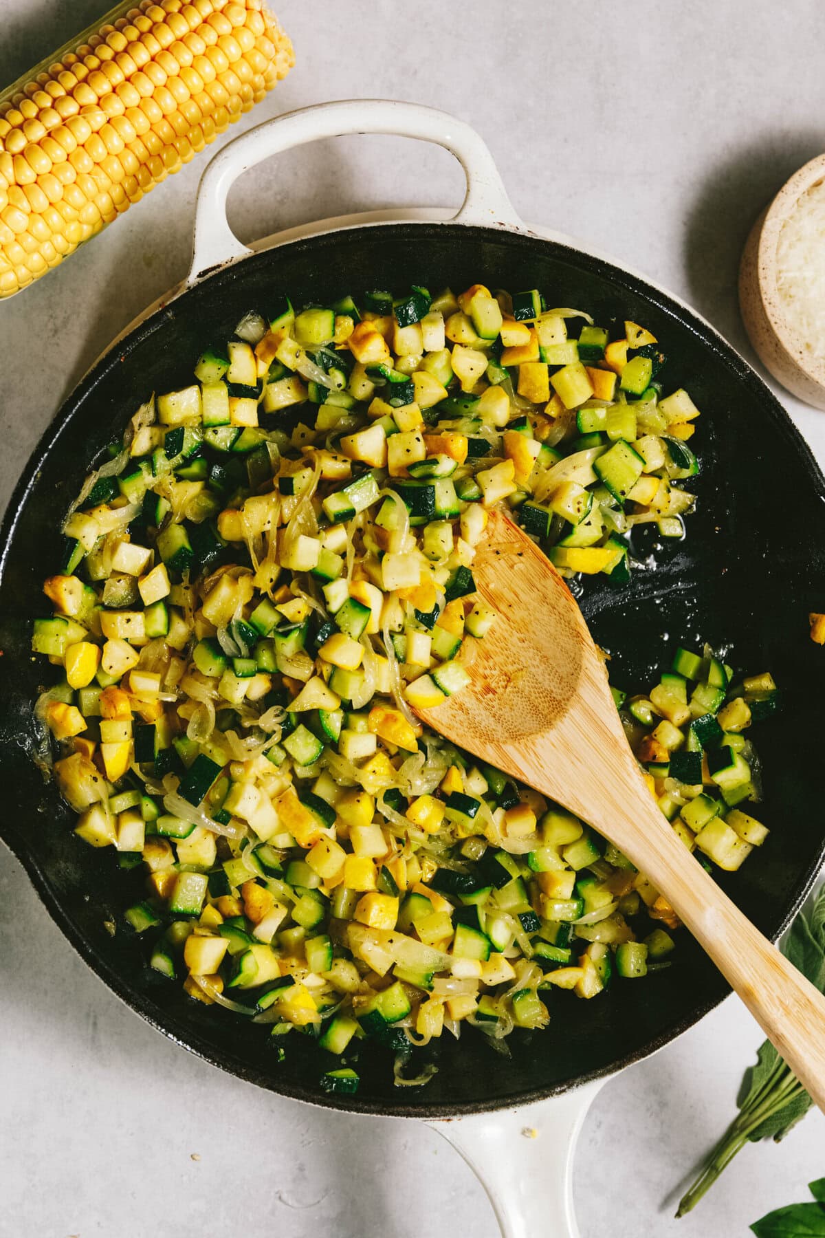 A skillet with sautéed diced zucchini, corn, and onions creates a vibrant mix reminiscent of summer squash pasta. A wooden spoon rests on the edge while fresh corn and a small bowl sit nearby on a light surface.