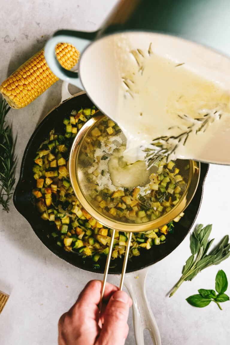 Pouring a creamy mixture through a sieve into a skillet with summer squash and other vibrant vegetables, next to a corn cob, rosemary, and basil leaves on a white surface, evokes the essence of an elegant summer squash pasta dish.