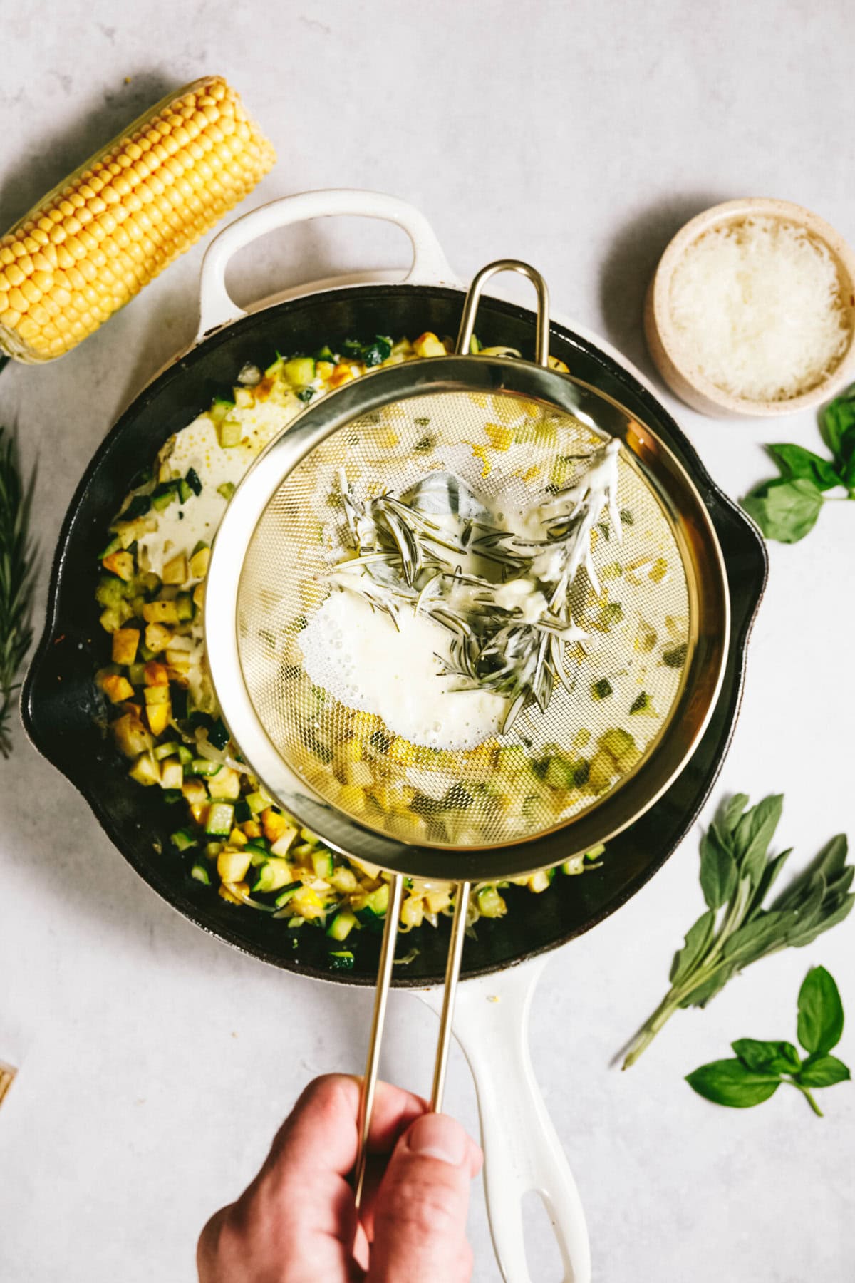 A hand holds a mesh strainer with herbs over a skillet containing diced vegetables and summer squash, ready to create a vibrant dish. Corn, cheese, and more fresh herbs await on the side, perfect for topping off your pasta masterpiece.