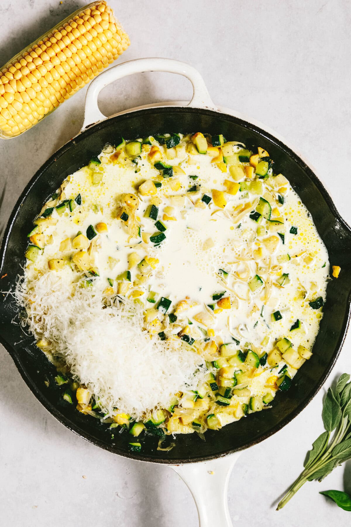 A skillet with a creamy mixture of diced zucchini and grated cheese sits beside a whole corn on the cob and a sprig of fresh herbs. It's reminiscent of summer squash pasta on a light surface.